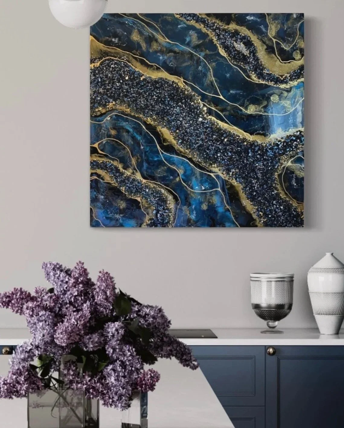 A modern indoor space featuring a large abstract painting on the wall with blue, black, gold, and white swirls and lines. A table with a bouquet of purple lilacs in a glass vase is in the foreground. On a blue sideboard are a decorative silver and bl