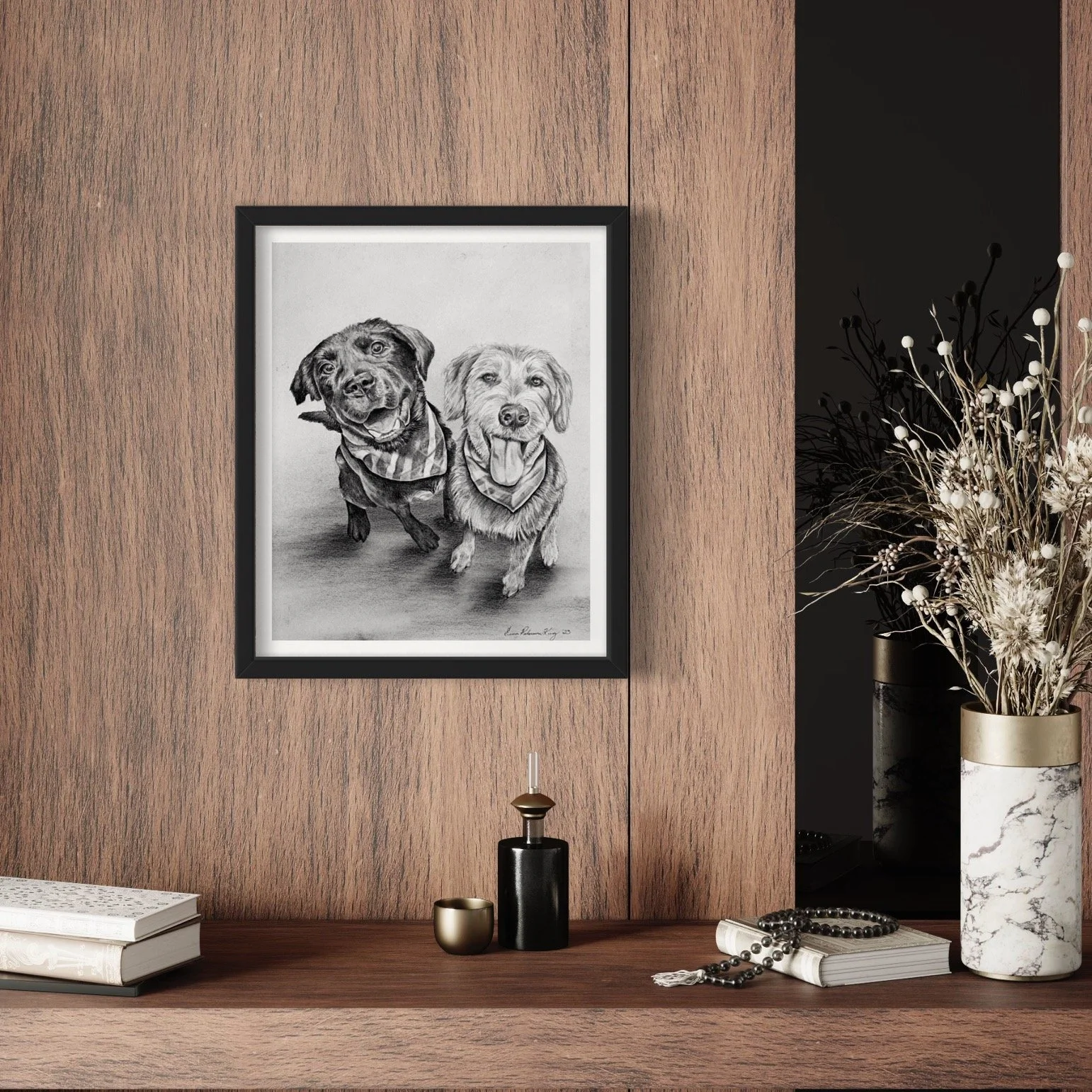 A wooden wall with a framed black and white drawing of two dogs, a black and tan one and a lighter one, both wearing bandanas. Below are black and gold-colored vases, a book, and a string of beads. To the right is a vase with dried white and black fl