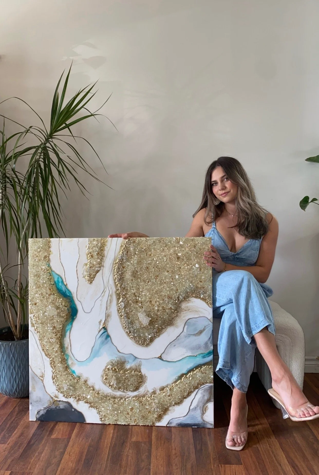 A woman sitting on a white chair, holding a large abstract art piece with gold, white, blue, and black textures, in a room with a hardwood floor and green plants.