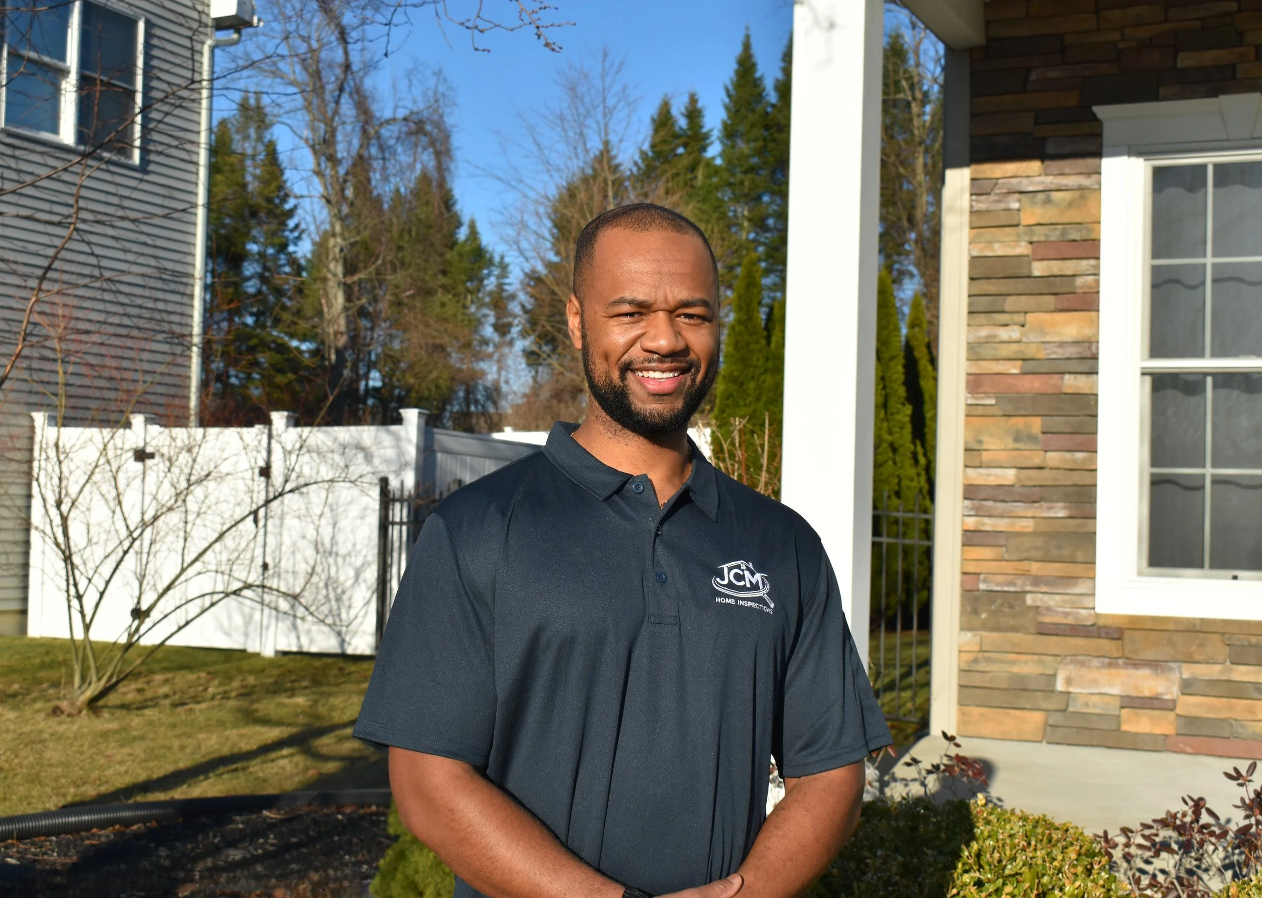 A man standing outdoors in front of a house, smiling, wearing a navy polo shirt with a company logo that says 'JCM Home Inspection', on a sunny day with a blue sky and trees in the background.