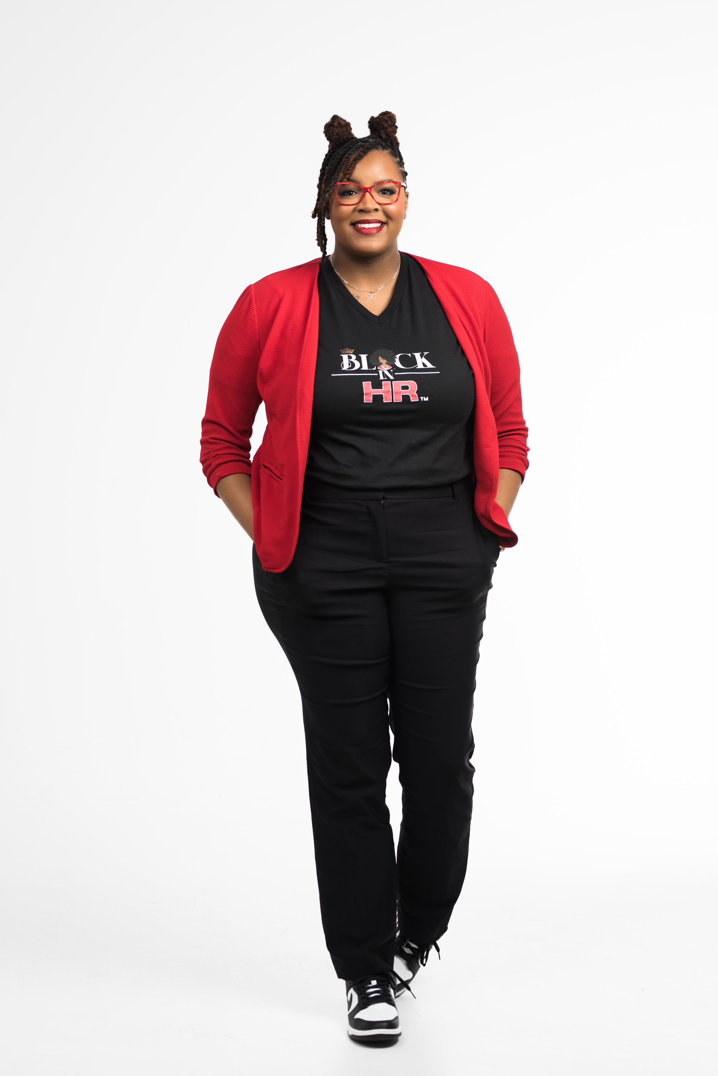 A woman with glasses and styled hair standing against a white background, wearing a black T-shirt with a logo and text, a red blazer, black pants, and sneakers, smiling with her hands in her pockets.