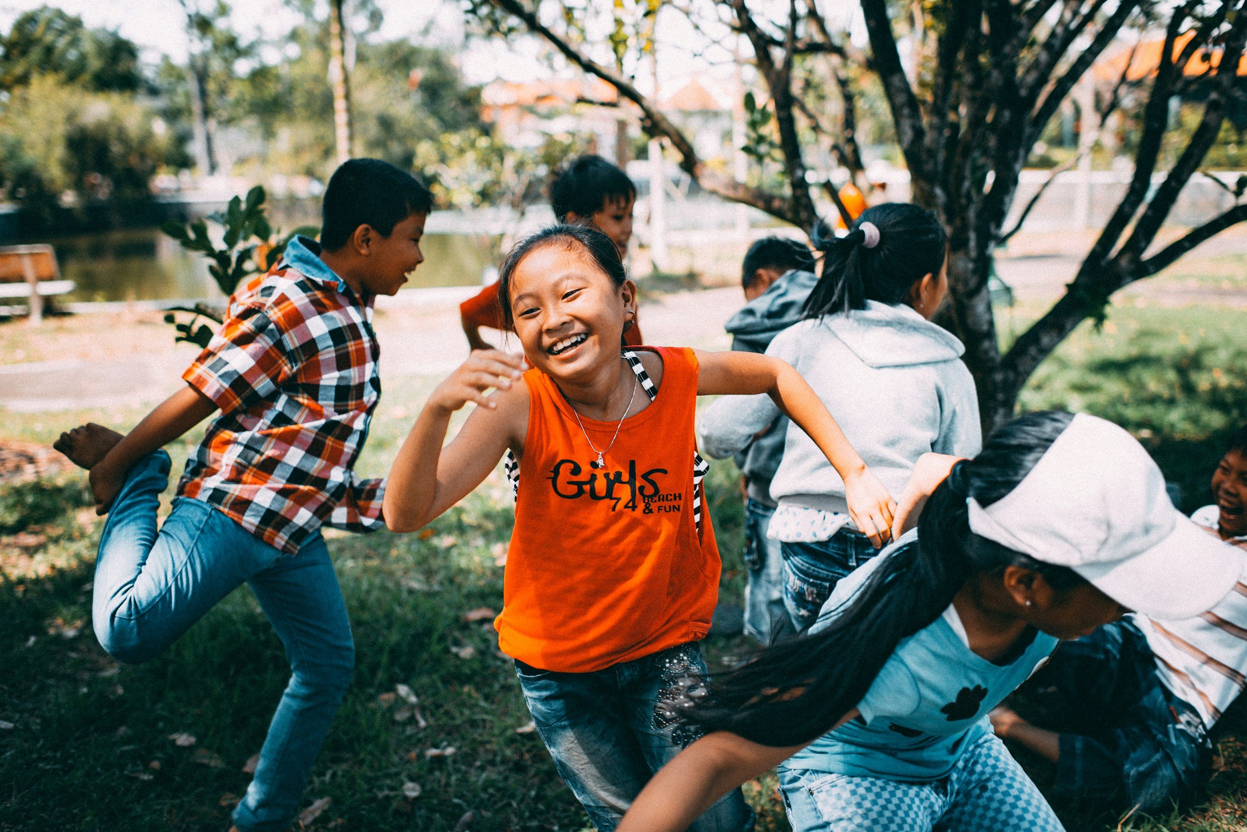 Children playing and laughing outdoors near a tree by a pond.