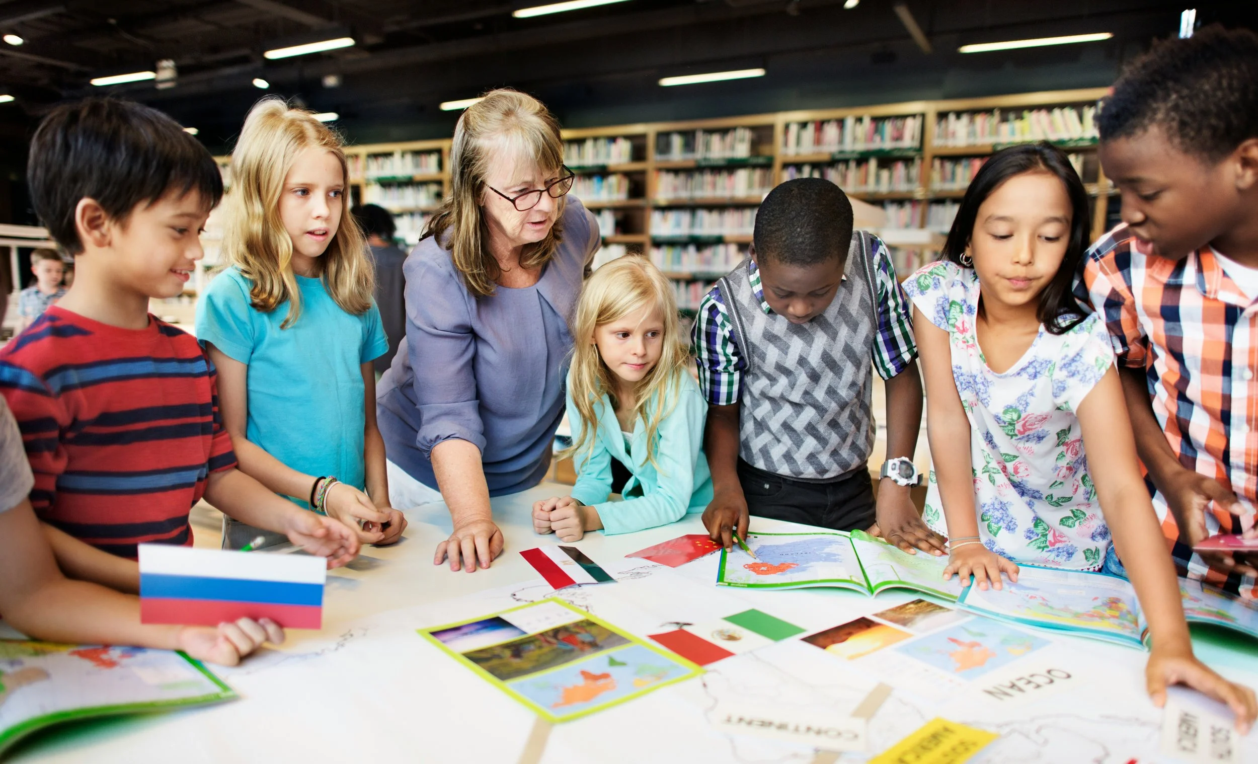 Group of diverse children and an elderly woman gathered around a table in a library, looking at books and maps with flags, engaging in a learning activity.