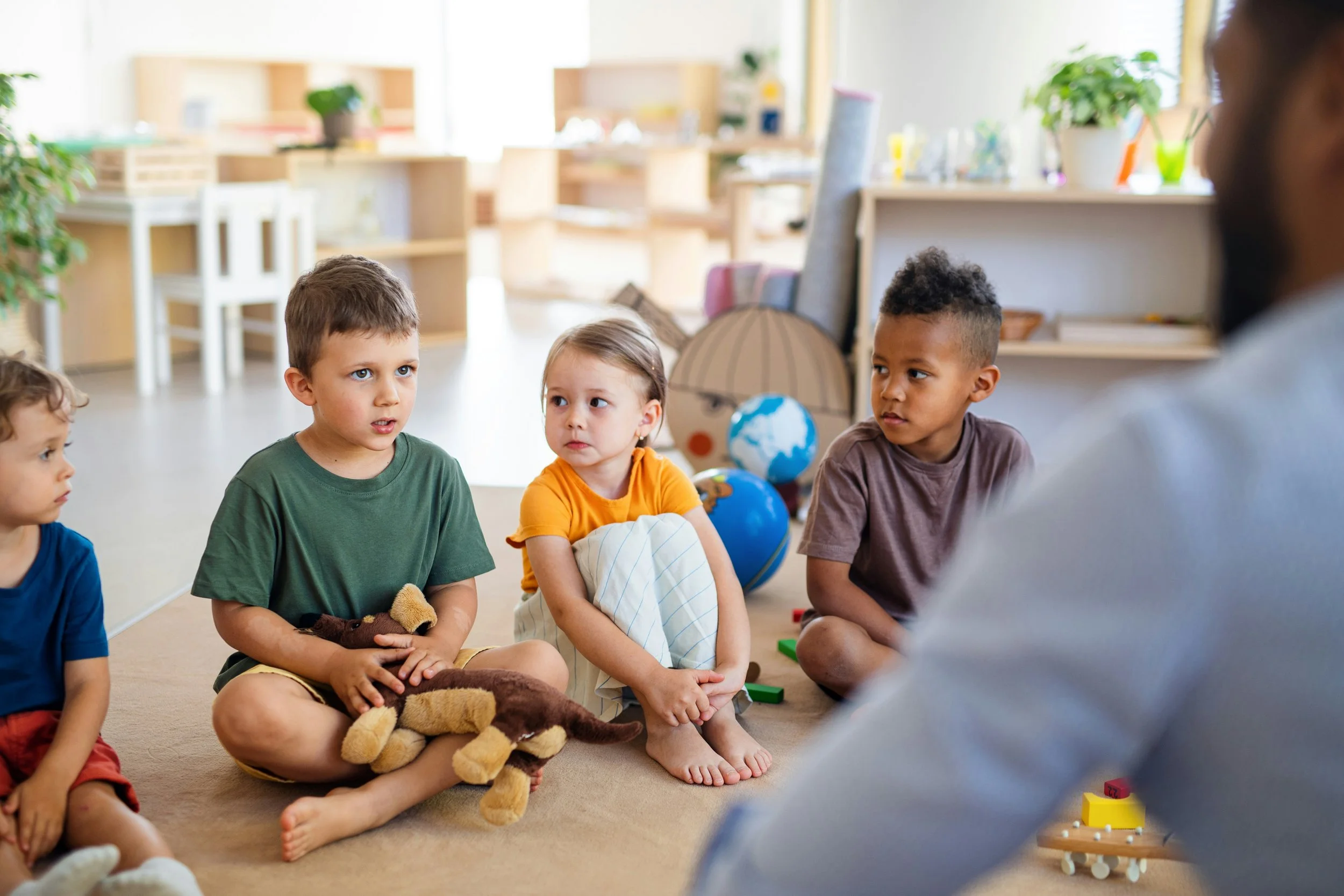 Four young children sitting on the floor in a classroom, listening to an adult. Classroom background includes toys, globes, and plants.