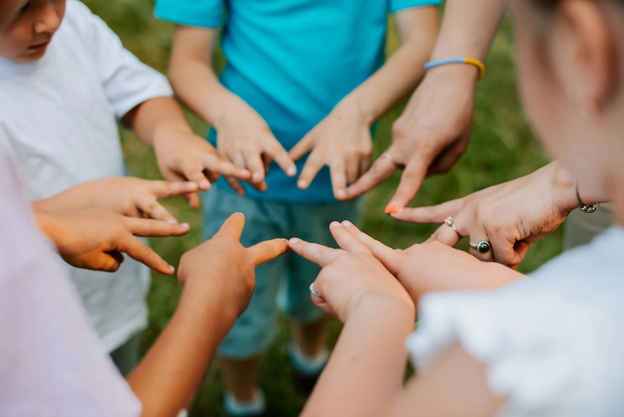 A group of children and adults with their hands stacked together in the center, outdoor setting on grass.
