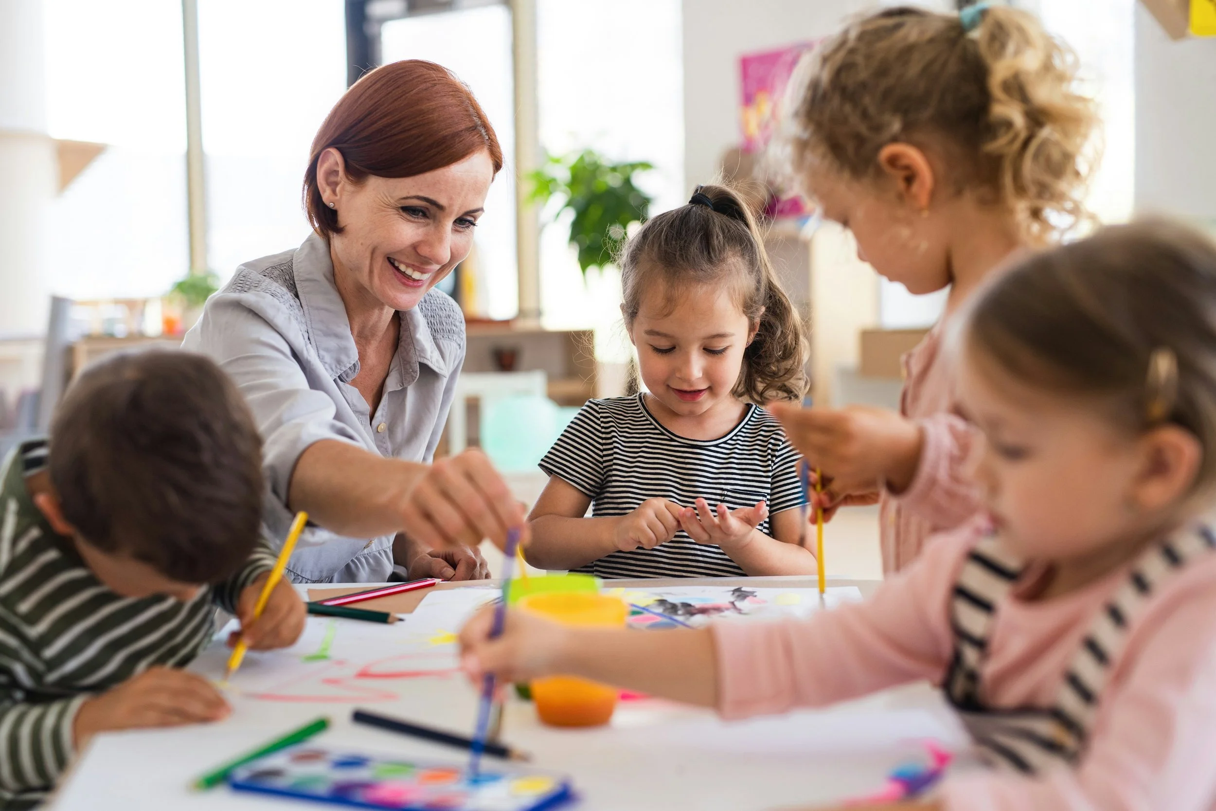 A woman and four children are engaged in a painting activity at a table in a bright room.