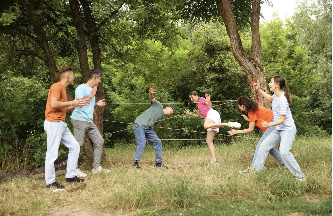 Group of six young people playing a game of tug-of-war outdoors in a wooded area.