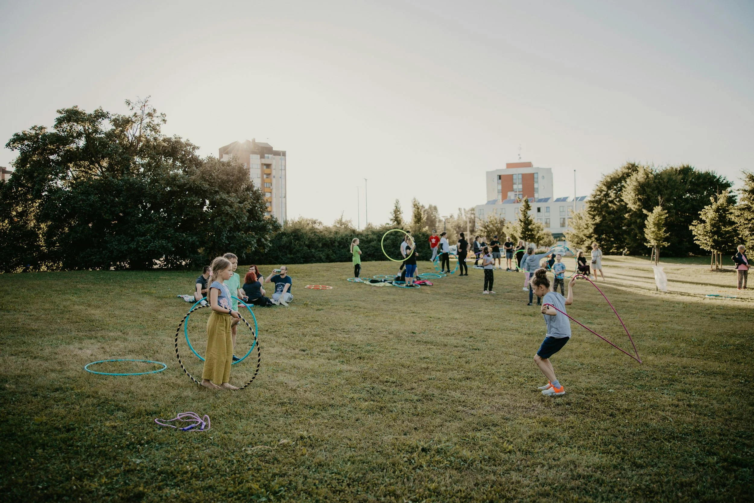 Children playing with hula hoops in a park during late afternoon or early evening, with adults sitting on the grass and city buildings in the background.
