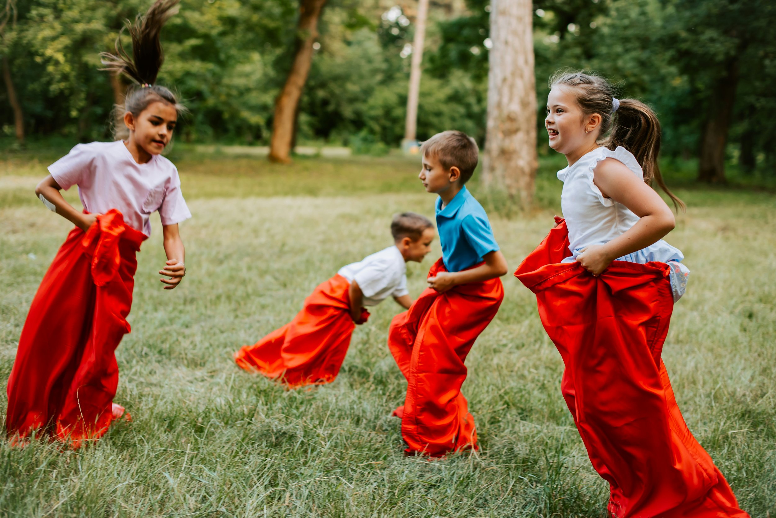 Children playing a sack race outdoors on grass in a wooded area.