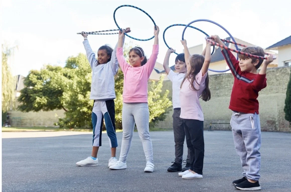 Five children playing outdoors on a paved surface, holding hula hoops above their heads. Trees and houses are visible in the background.