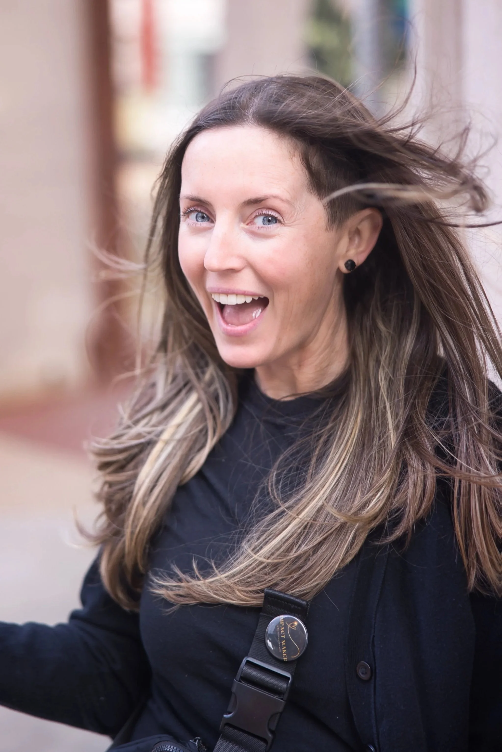 A woman with long, flowing hair, smiling with an open mouth, outdoors with blurred buildings in the background.