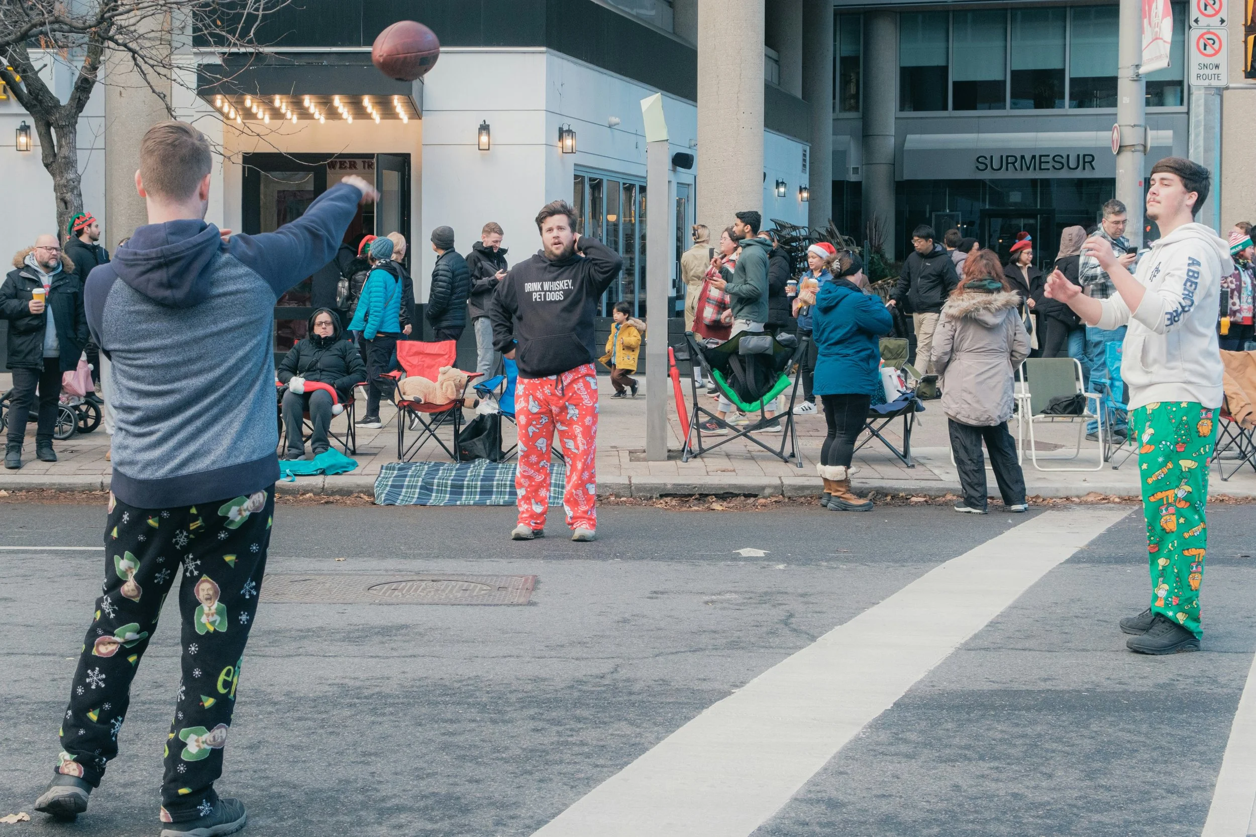 Three men in pajamas playing football on a city street during a Christmas celebration with people watching from the sidewalk and tents set up.