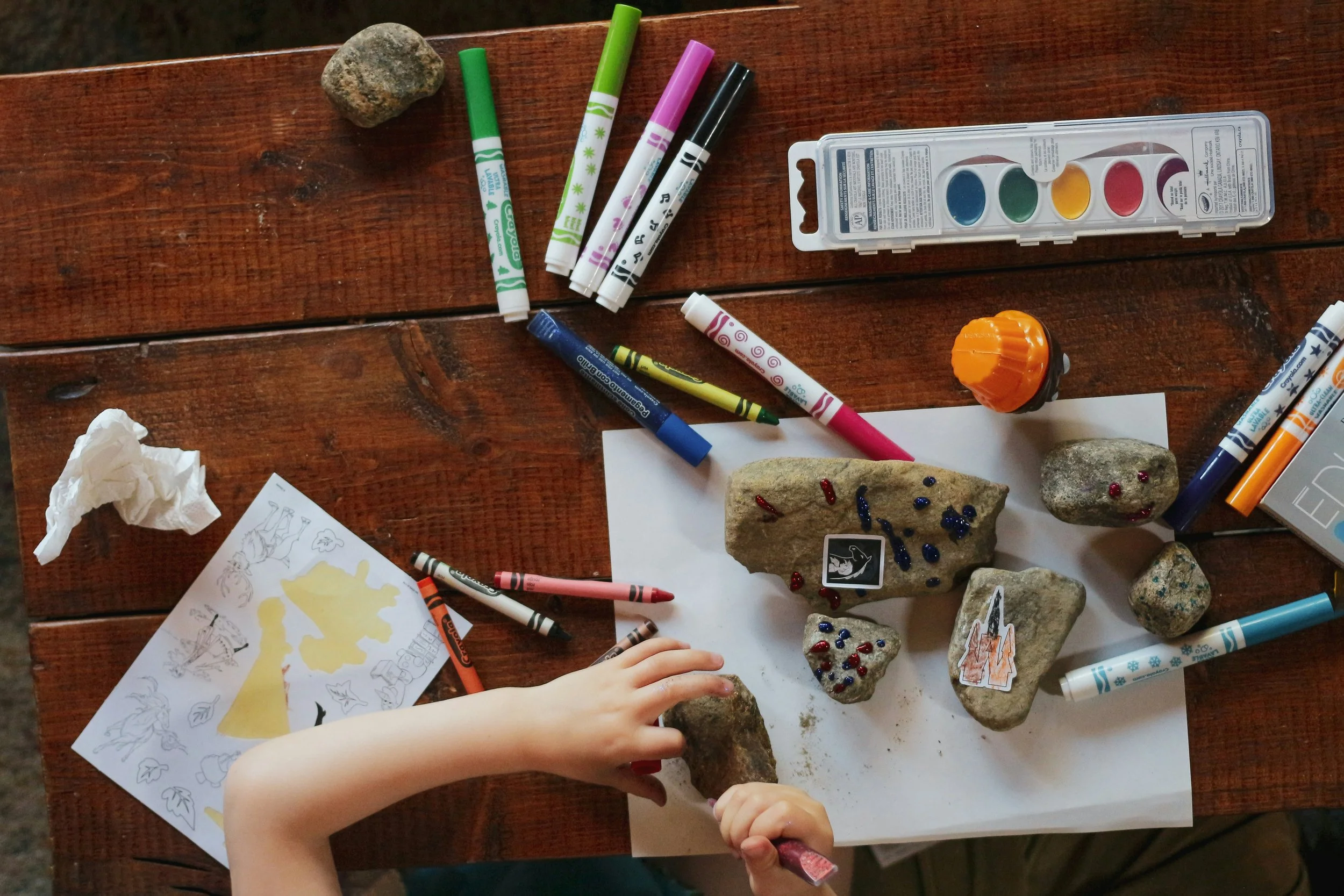 Child's hand reaching to place a rock on a white sheet surrounded by various larger rocks decorated with small red and blue beads, markers of different colors, a crumpled tissue, a coloring sheet with outdoor scenes, and watercolor paints in a plastic case on a wooden table.