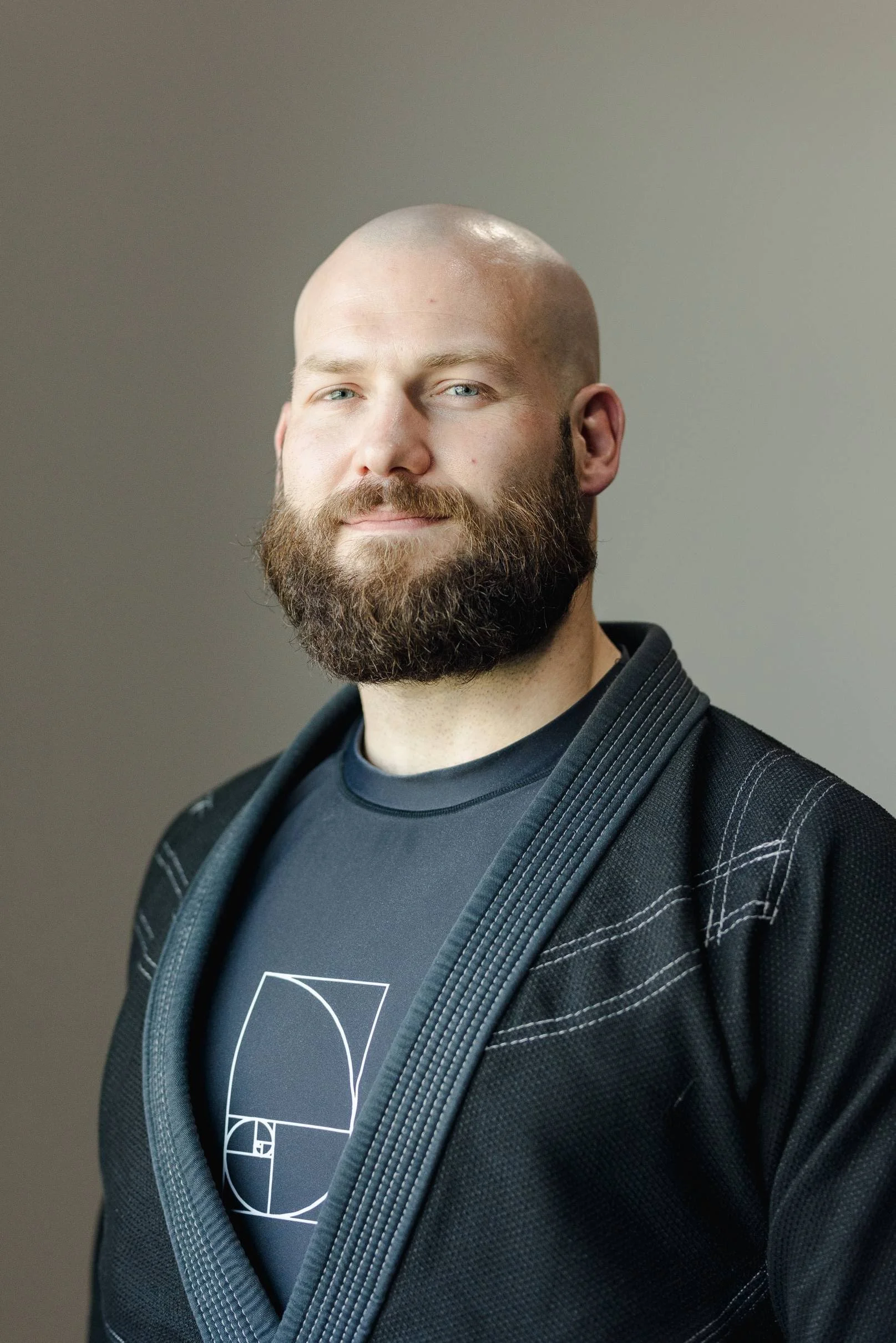 A man with a shaved head and a full beard, wearing a black martial arts gi with white stitching and a black t-shirt featuring a white geometric Fibonacci spiral design.