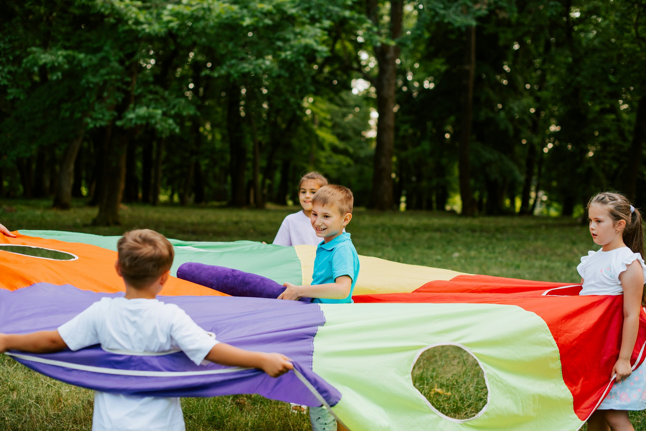 Children playing with a colorful parachute outdoors in a park surrounded by trees.