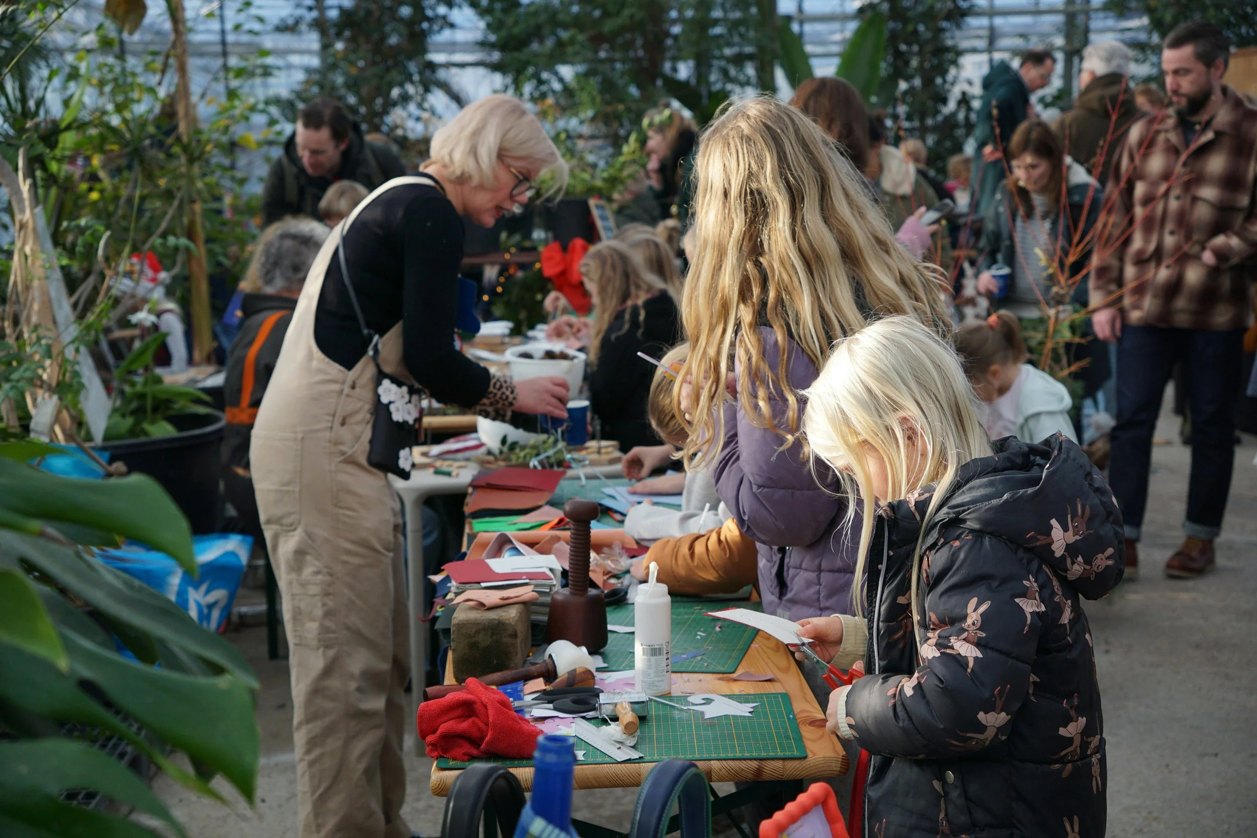 People gathered at a table engaging in crafting activities inside a greenhouse surrounded by green plants.