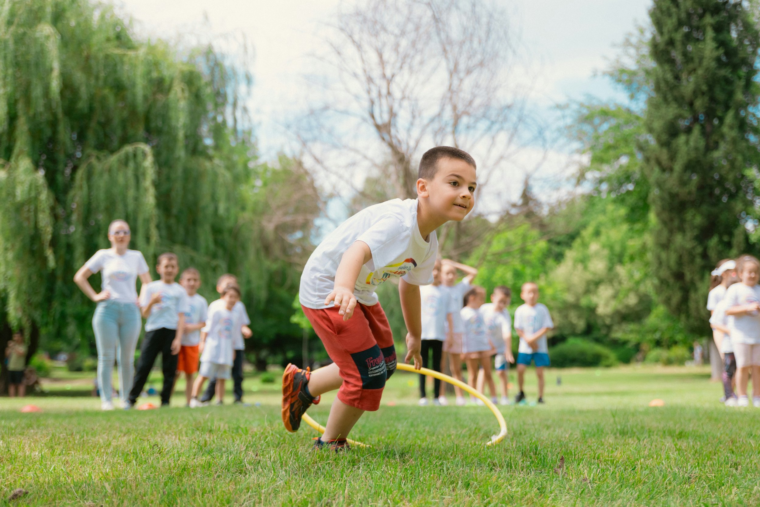 A young boy jumps through a hula hoop during an outdoor activity with a group of children and adults in a park with trees.