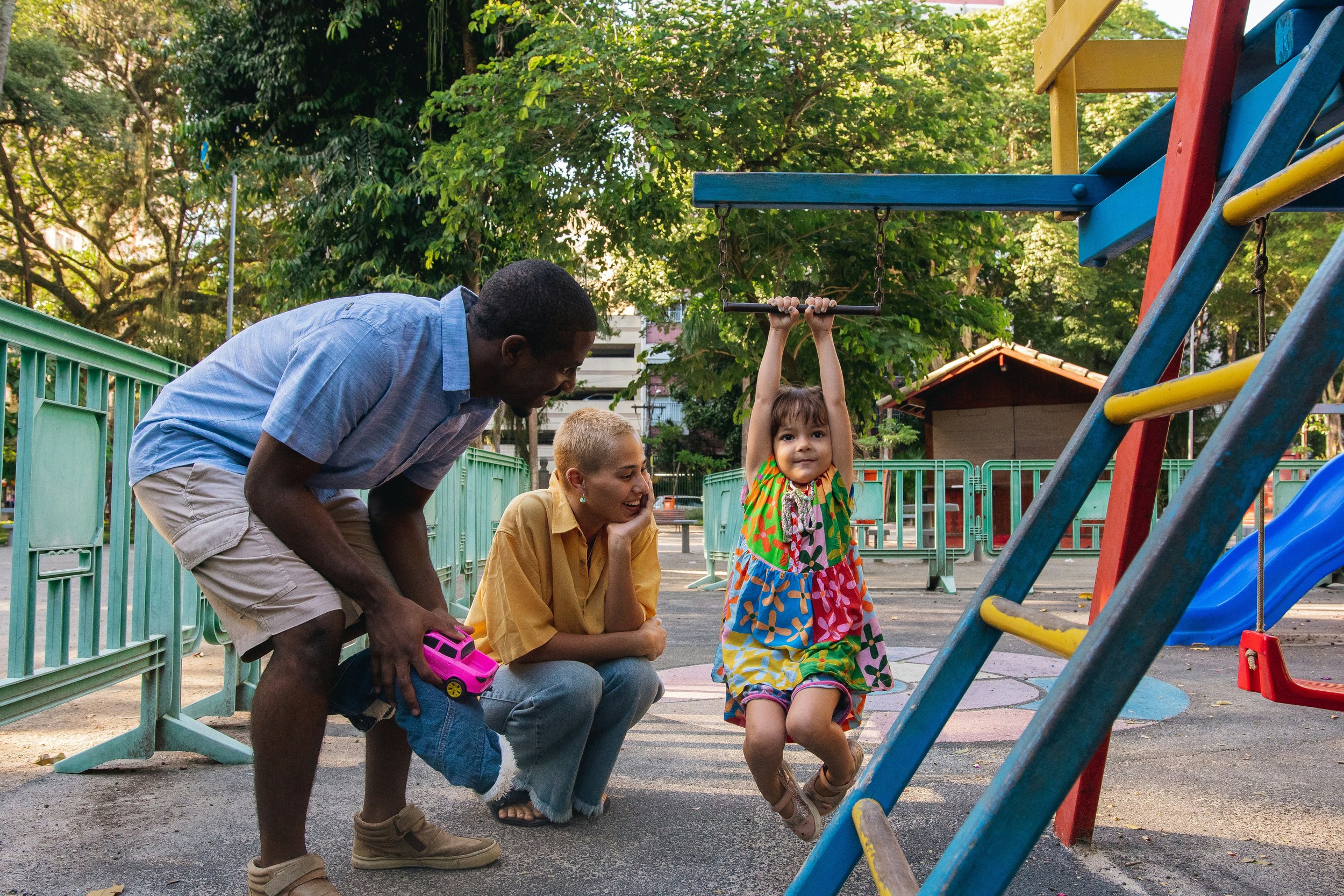 A young girl in a colorful dress hanging on a swing at a playground, with a woman and a man, likely her guardians, watching and smiling nearby. The scene is outdoors with trees and playground equipment.