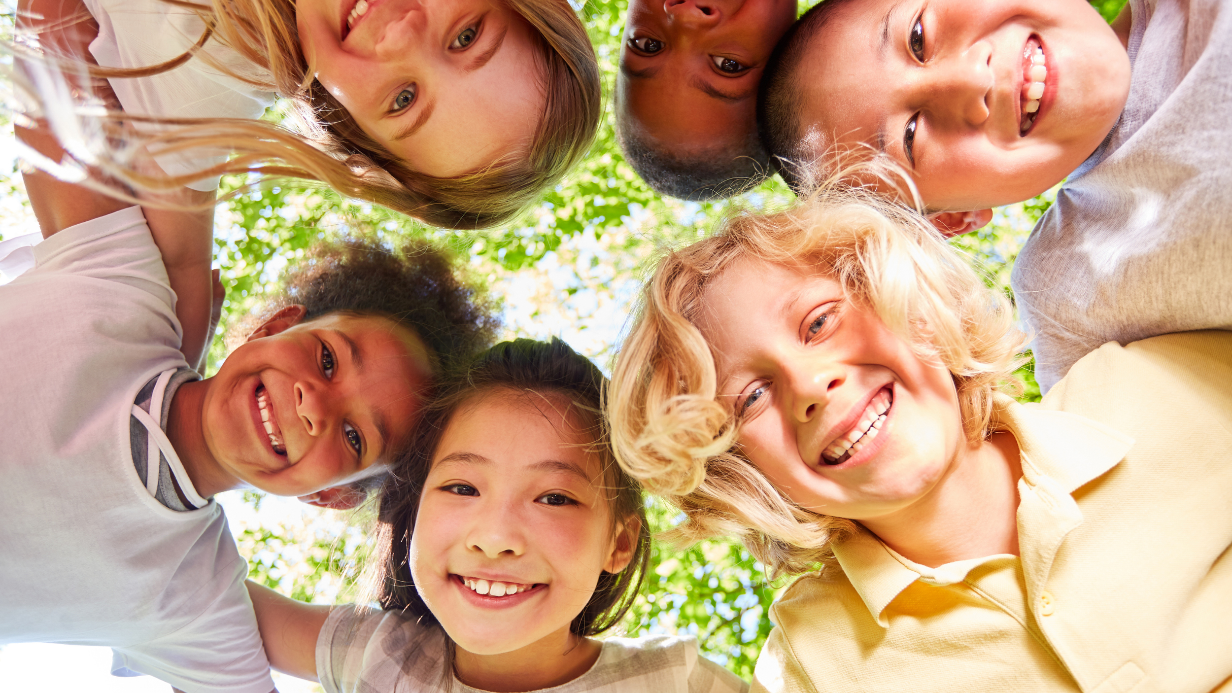A diverse group of children looking down into the camera, smiling and forming a circle outdoors under green trees.