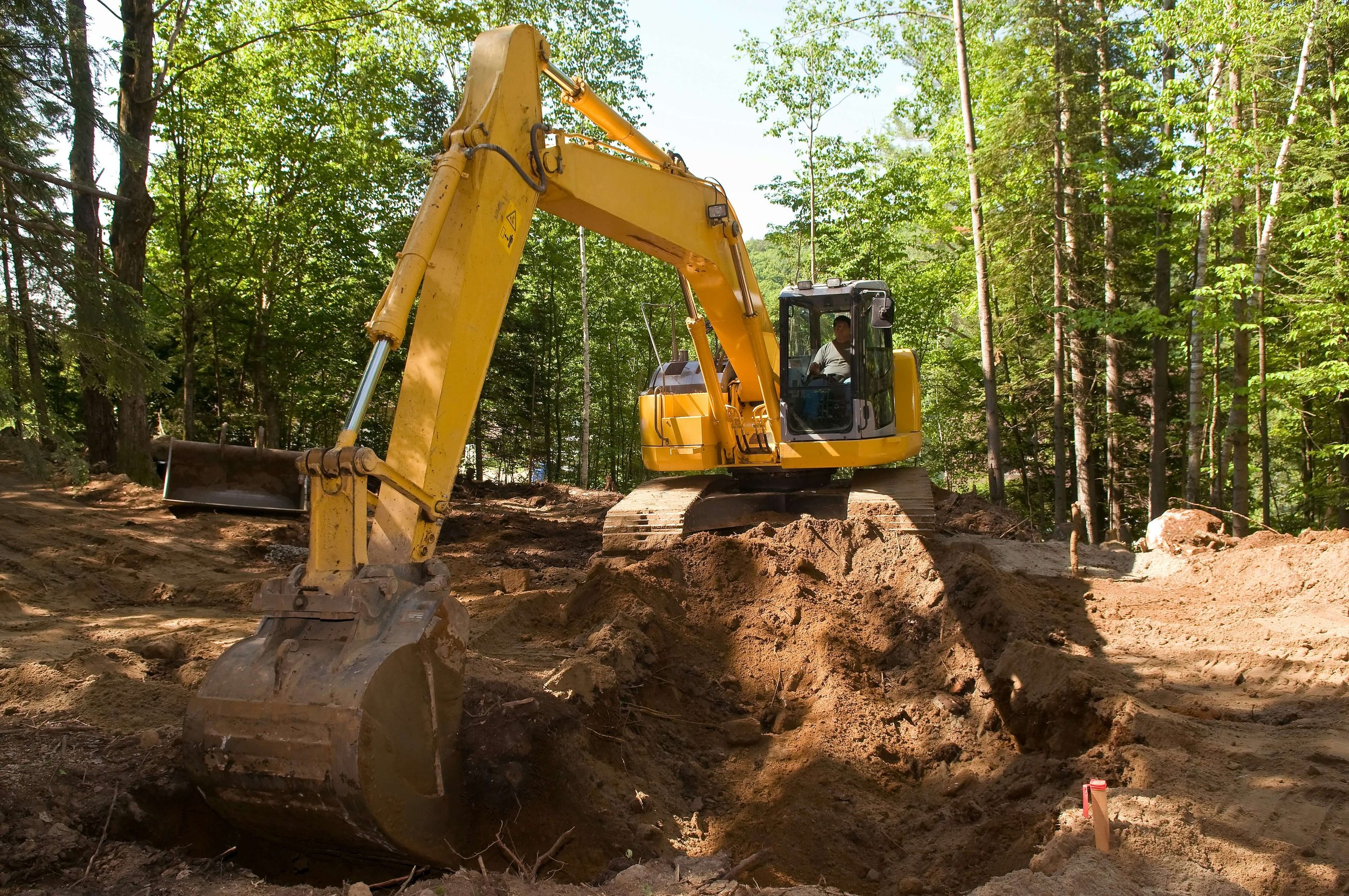 A yellow excavator with Alabama Landworks digging a trench in a forested area with green trees and dirt to perform land clearing services.
