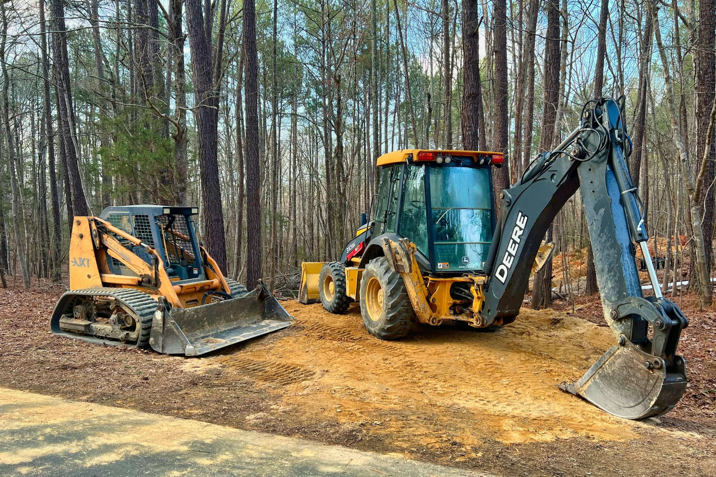 Construction equipment, including an excavator and a smaller machine, working on a dirt patch in a wooded area with tall trees and blue sky in the background with Alabama Landworks for land development services in Alabama.