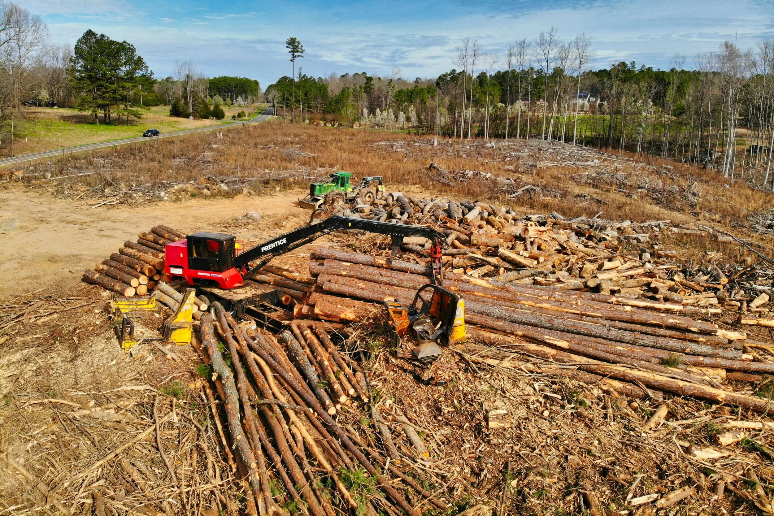 A scene of a tree and land clearing site for Alabama Landwork Services with logs and a wood chipper in the foreground, and a rural landscape with trees, a road, and houses in the background.