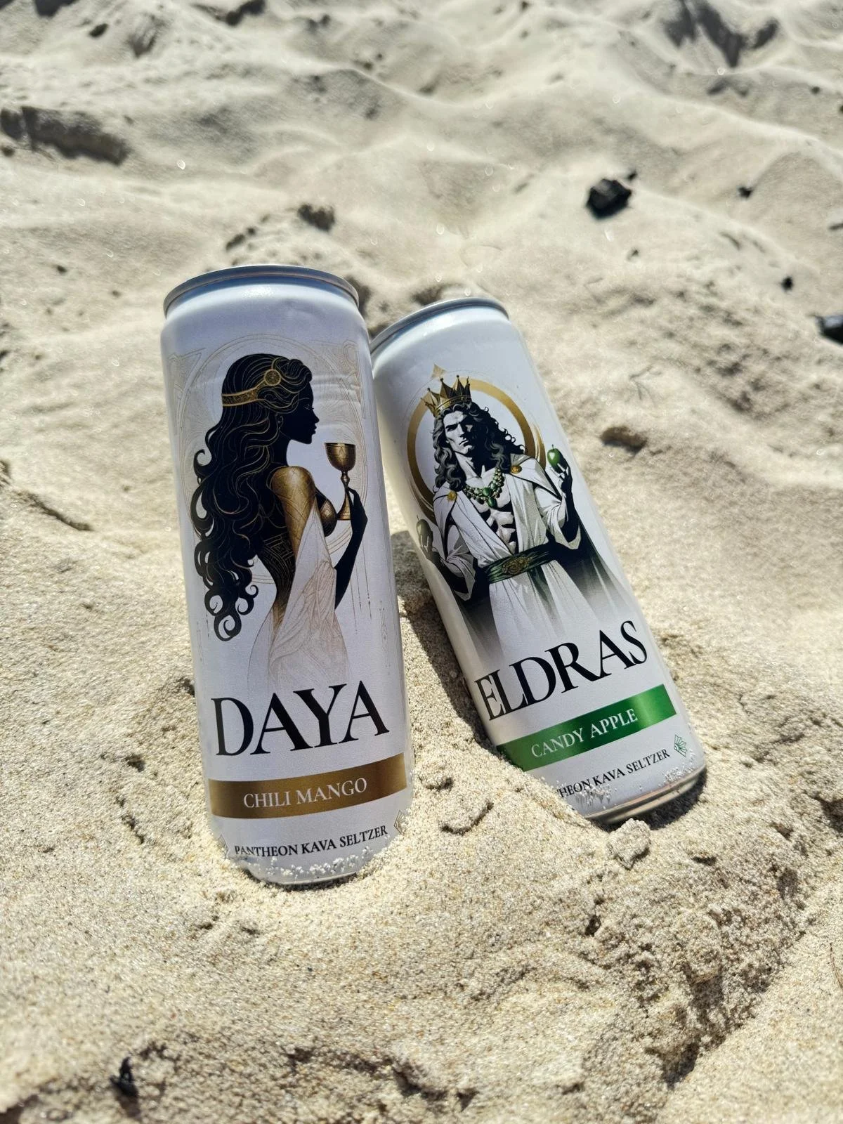 Two cans of elixirs with fantasy-themed labels featuring a woman and a king, placed on sandy beach. One labeled 'DAYA' with 'Chili Mango' flavor colored gold, and the other labeled 'ELDRAS' with 'Candy Apple' flavor colored green.