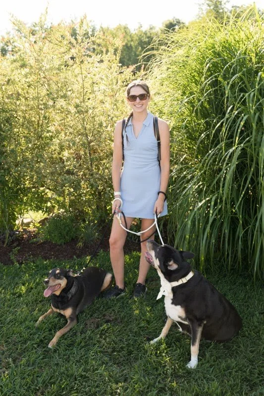 A young woman in a light blue dress and sunglasses standing on grass with two dogs, one sitting and one lying down, surrounded by green bushes and trees.