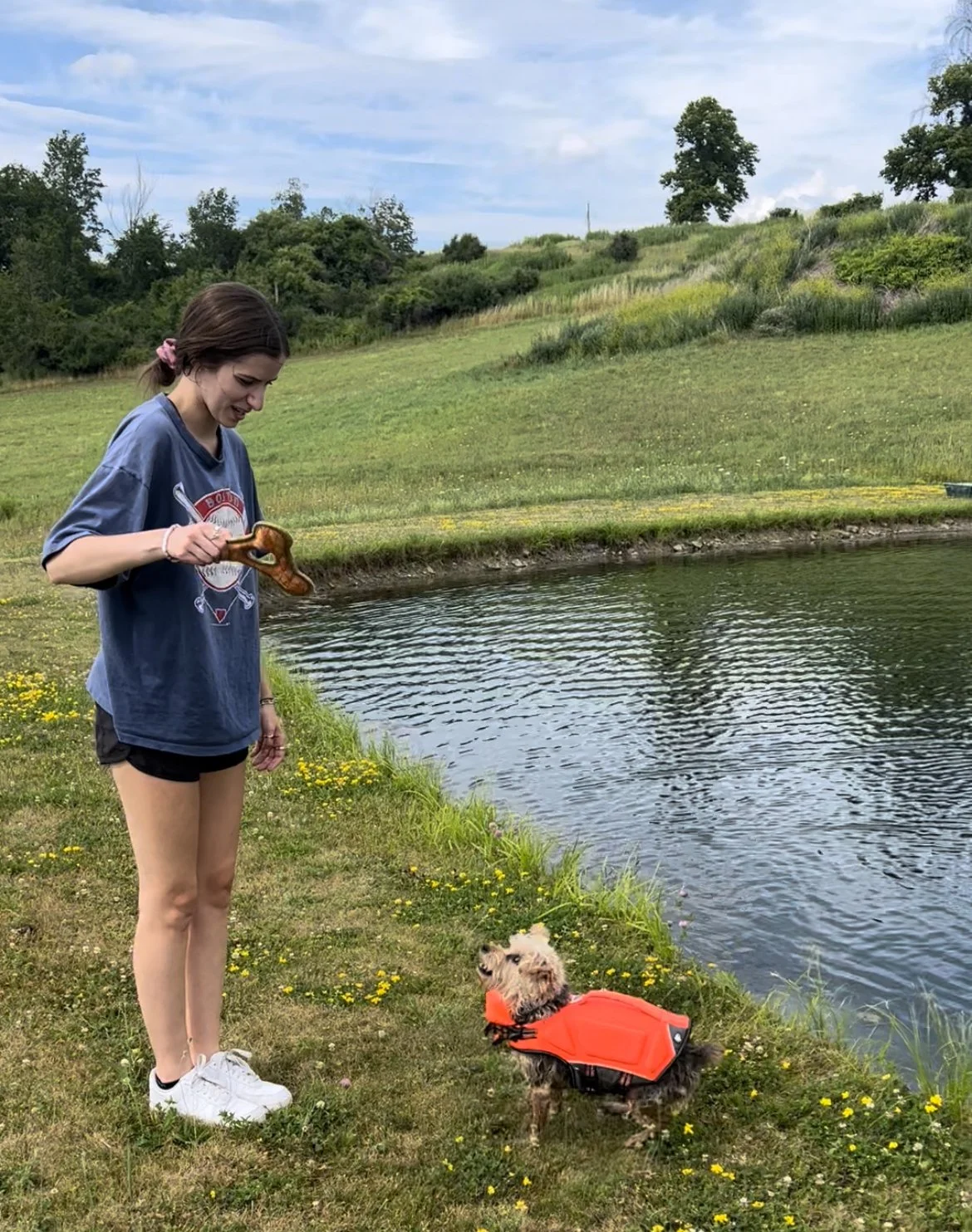 Liz standing next to a small Yorkie Poodle mix dog in an orange life jacket by a pond in a green field with trees and hills in the background.