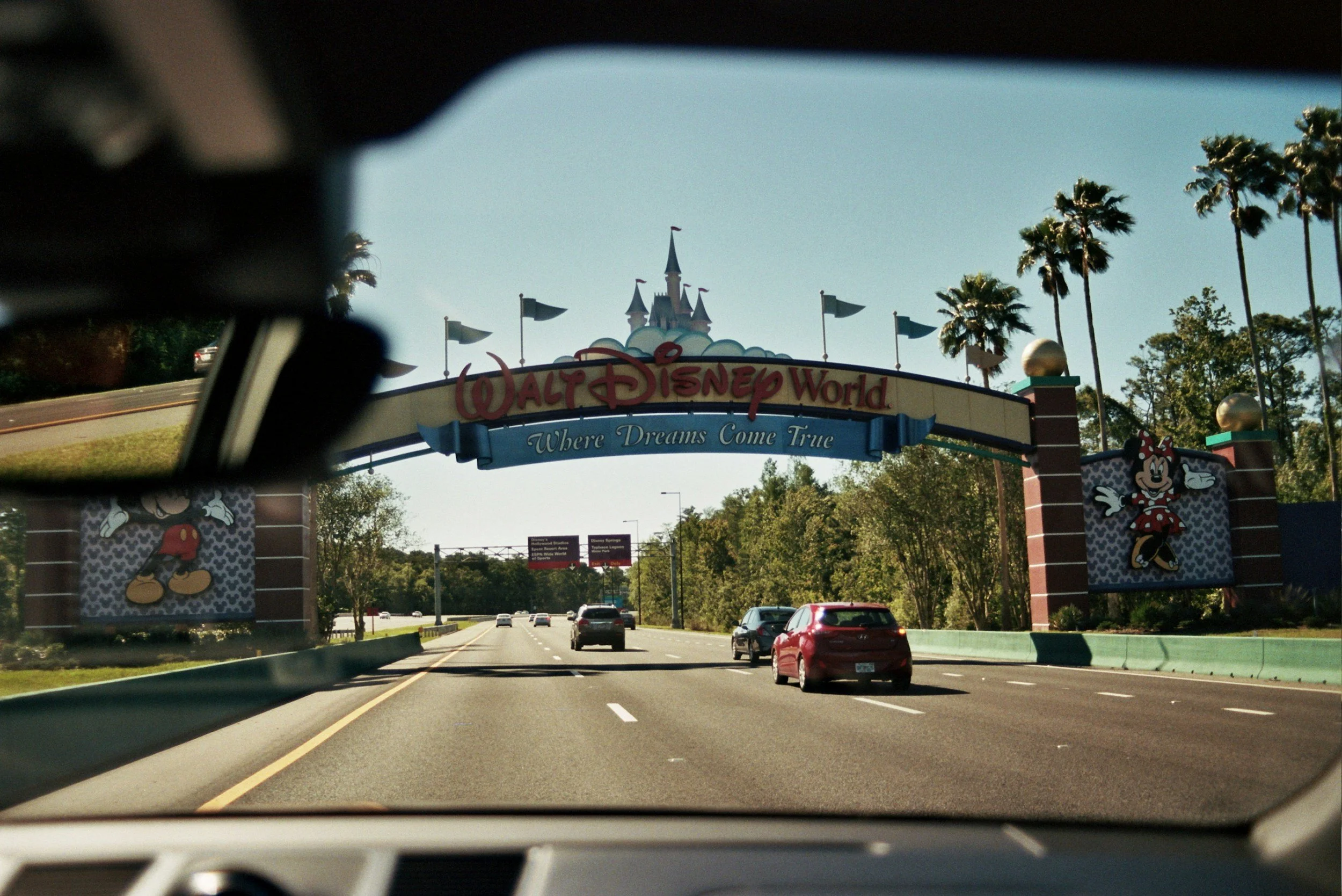 Entrance sign for Walt Disney World with castle silhouette, surrounded by palm trees, seen from inside a vehicle on a road.