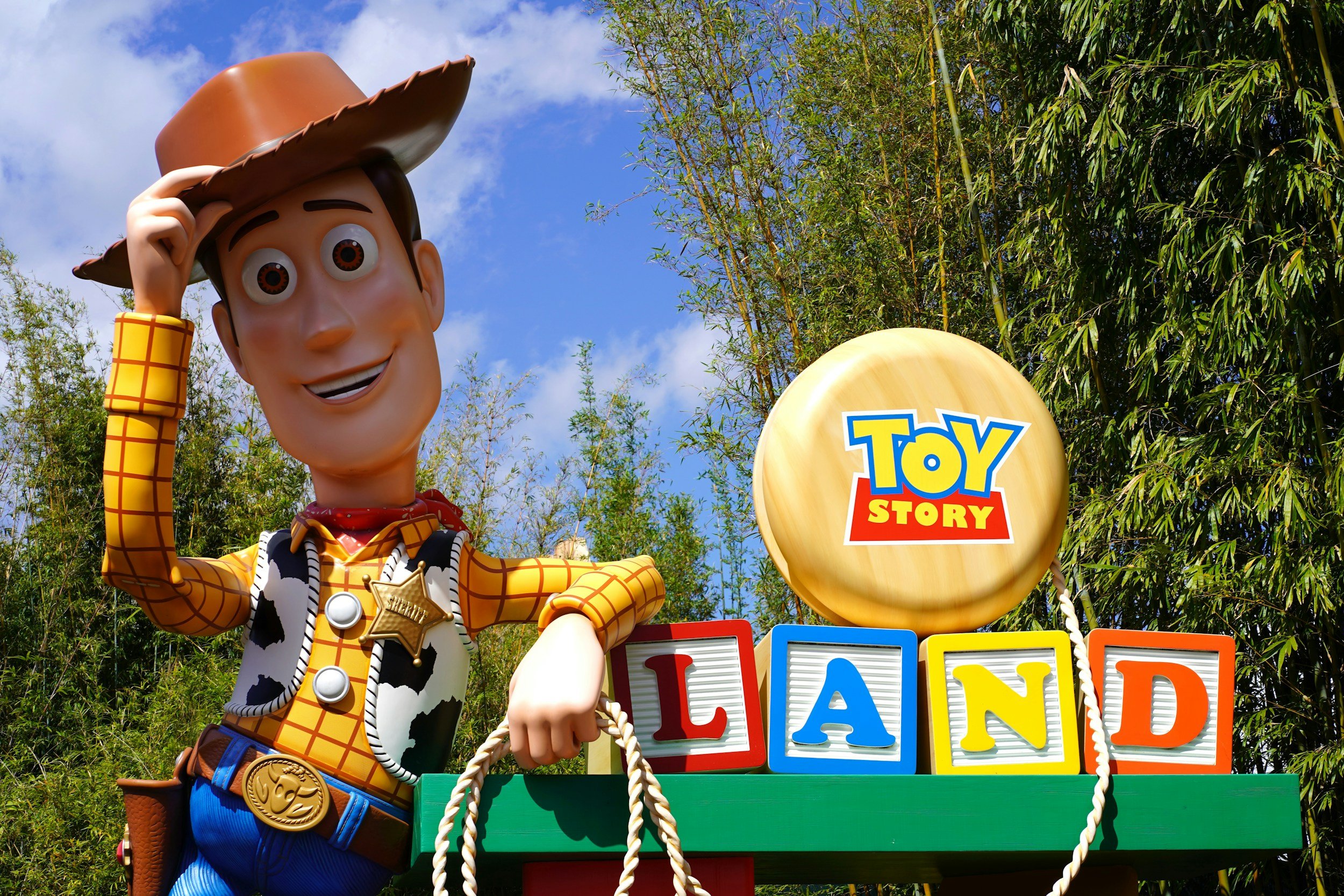 A life-sized figure of Sheriff Woody from Toy Story is adjusting his cowboy hat in front of colorful blocks spelling out 'LAND' and a round sign with the Toy Story logo. Green trees and a blue sky with clouds are in the background.