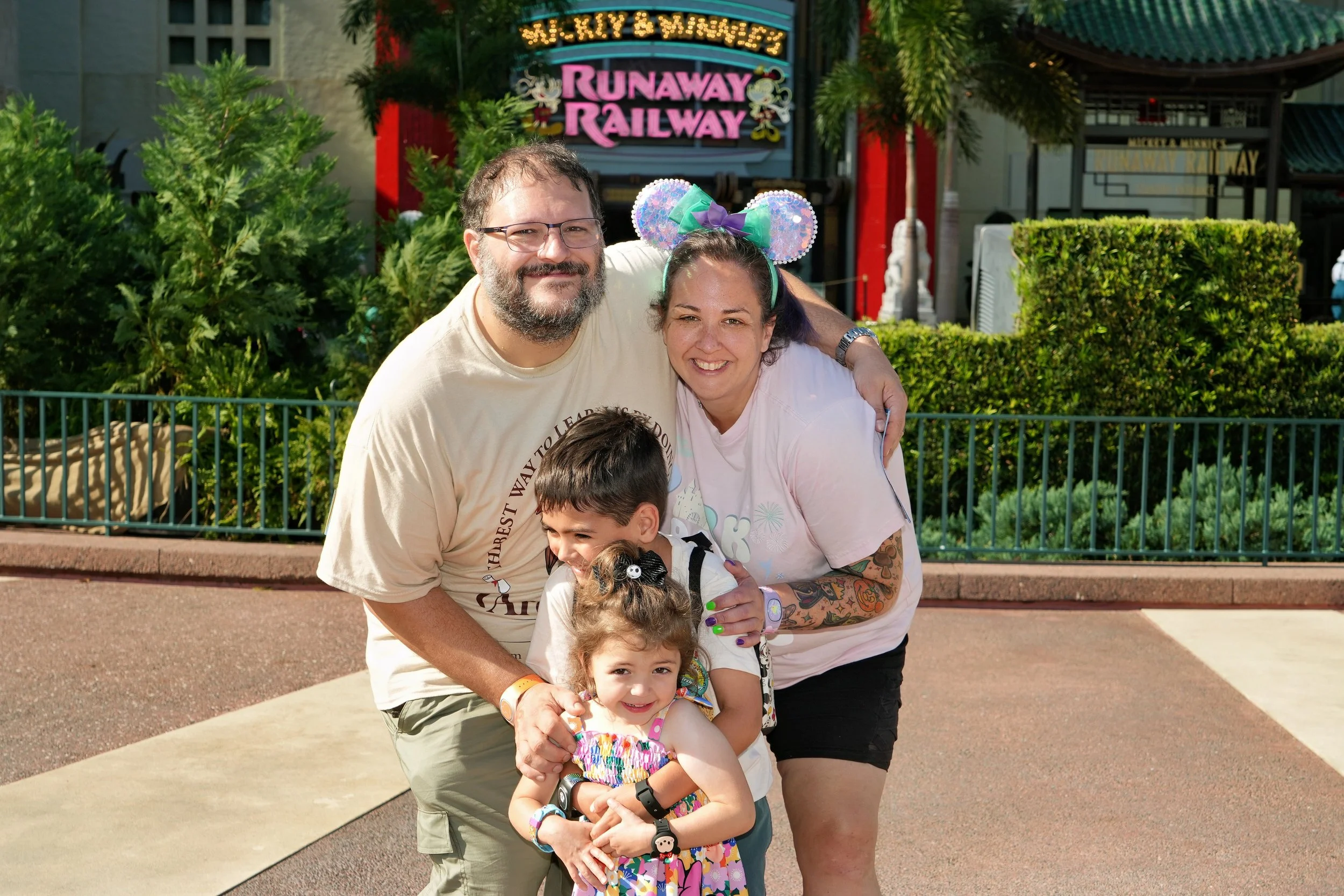A group of five people, including two adults and three children, smiling and posing together outside at a theme park. They are standing on a sidewalk with lush greenery and a fence behind them. In the background, there are colorful signs and themed structures, including one that says 'Runaway Railway,' indicating they are at a Disney park.