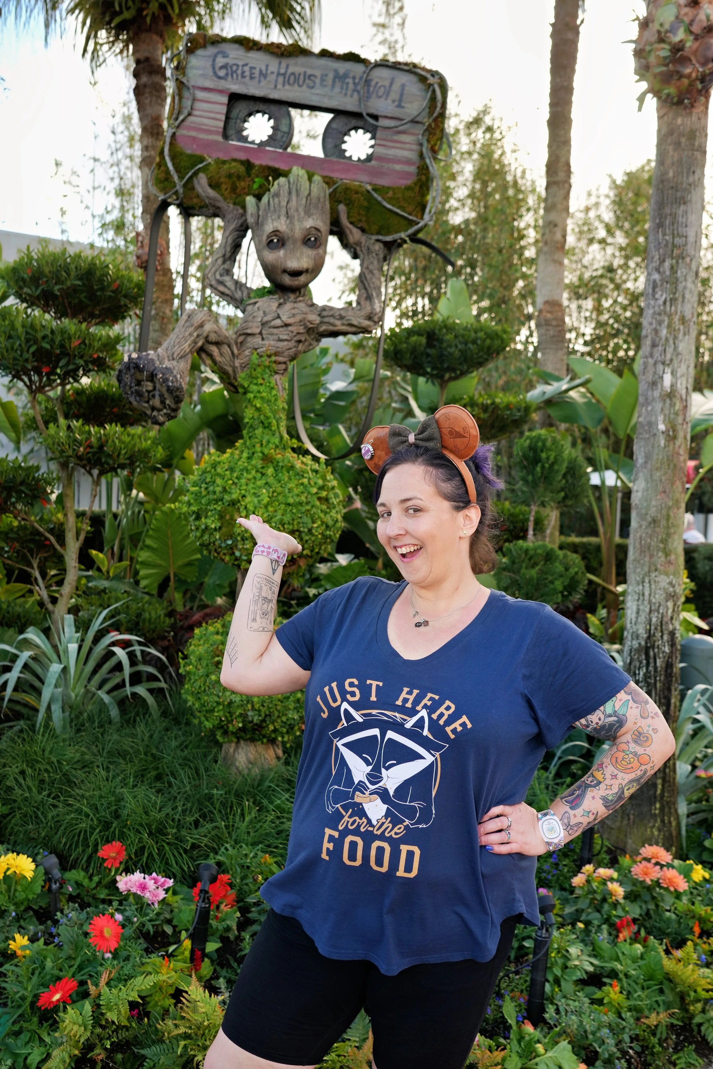 A young woman with tattoos on her right arm and wearing a Star Wars themed shirt, black shorts, and a Disney parks headband with ears, stands in a colorful garden with flowers and green plants, pointing to a large Groot sculpture holding a sign that reads 'Greenhouse Max Vol 1'.