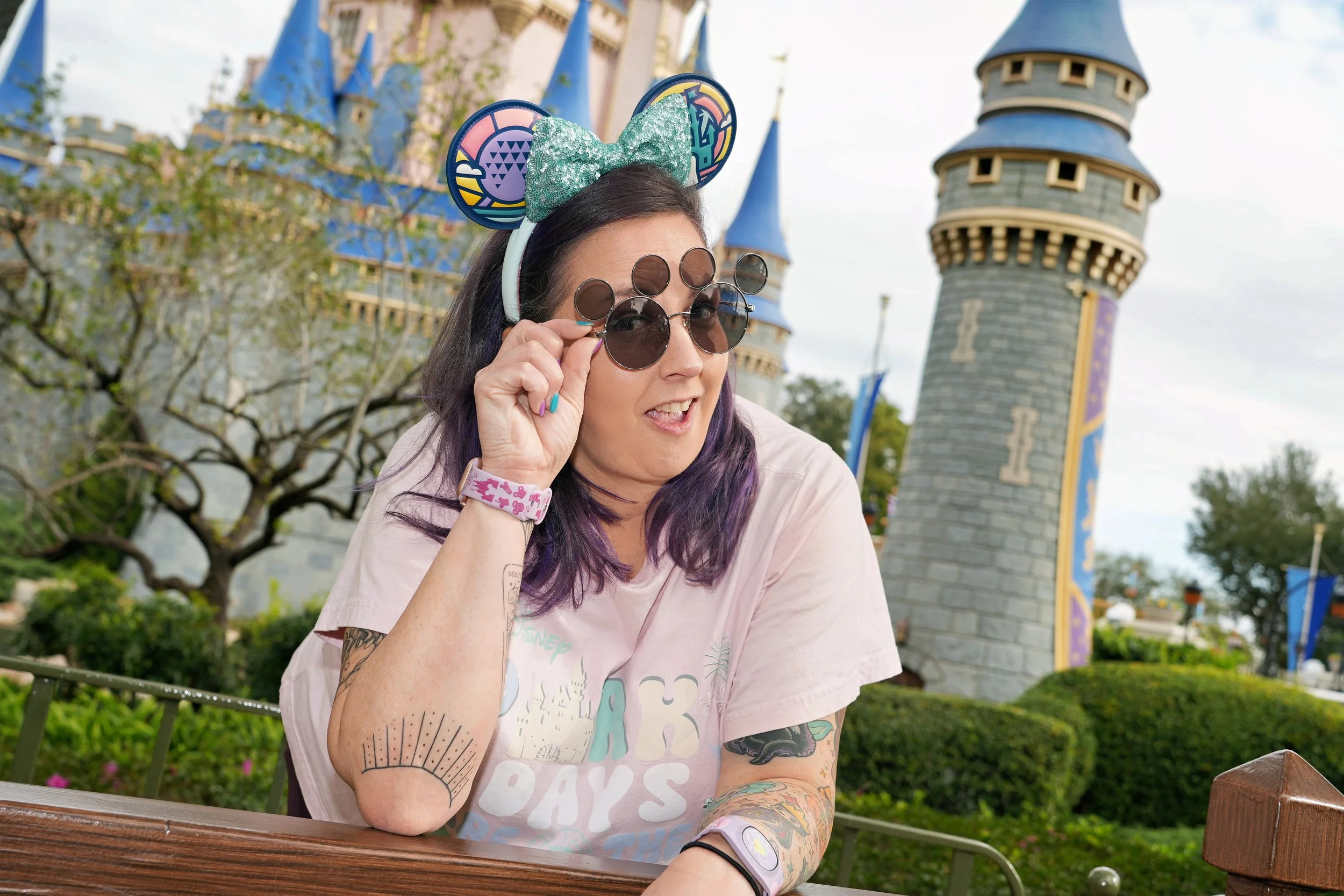 A woman with colorful tattoos on her arms wearing Disney ears with a glitter bow, sunglasses, and a pastel-colored T-shirt, sitting outdoors at Disneyland with the Sleeping Beauty Castle in the background.