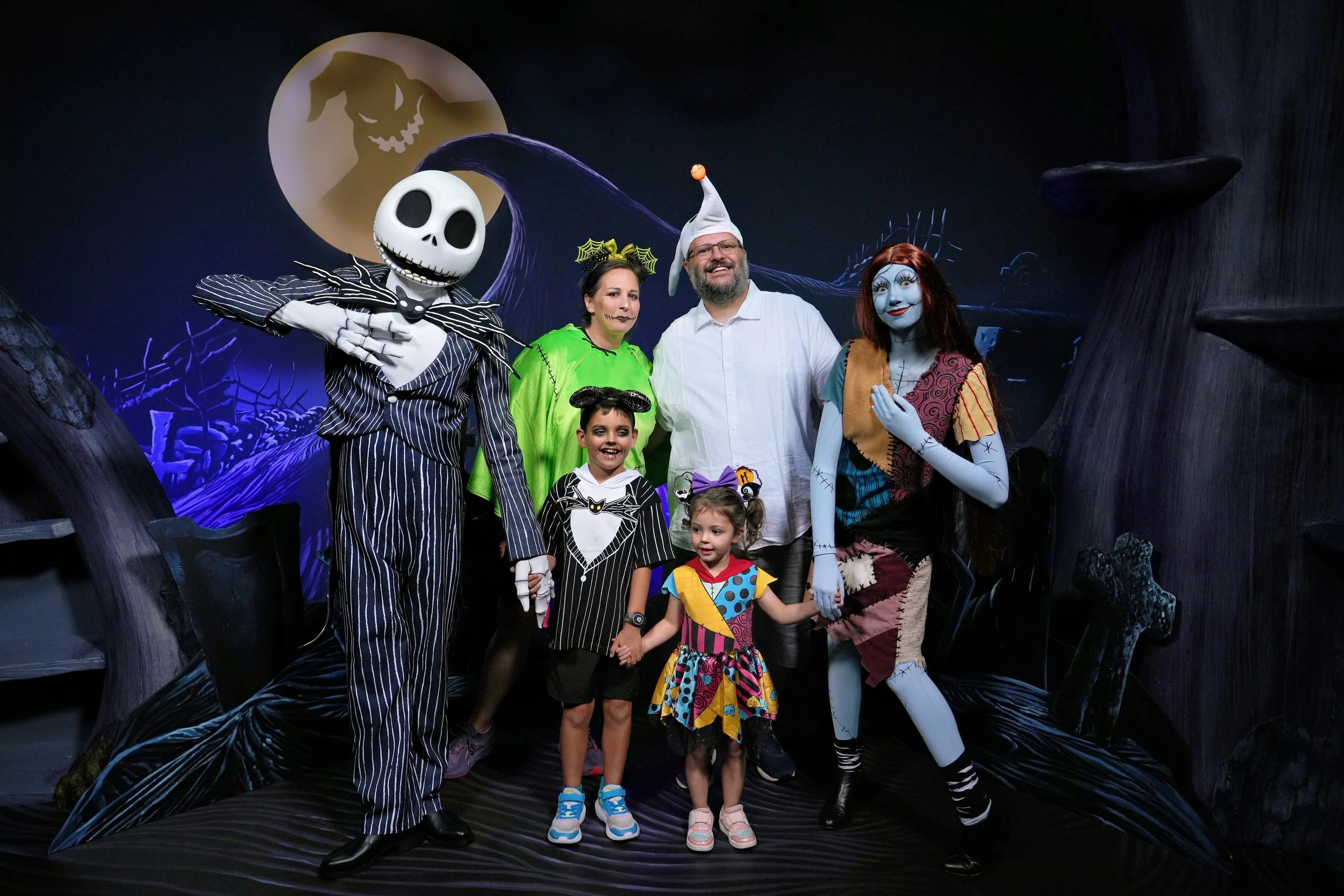 People dressed in Halloween costumes posing together in front of a spooky backdrop with a glowing moon and ghostly face, including characters like Jack Skellington from "The Nightmare Before Christmas."
