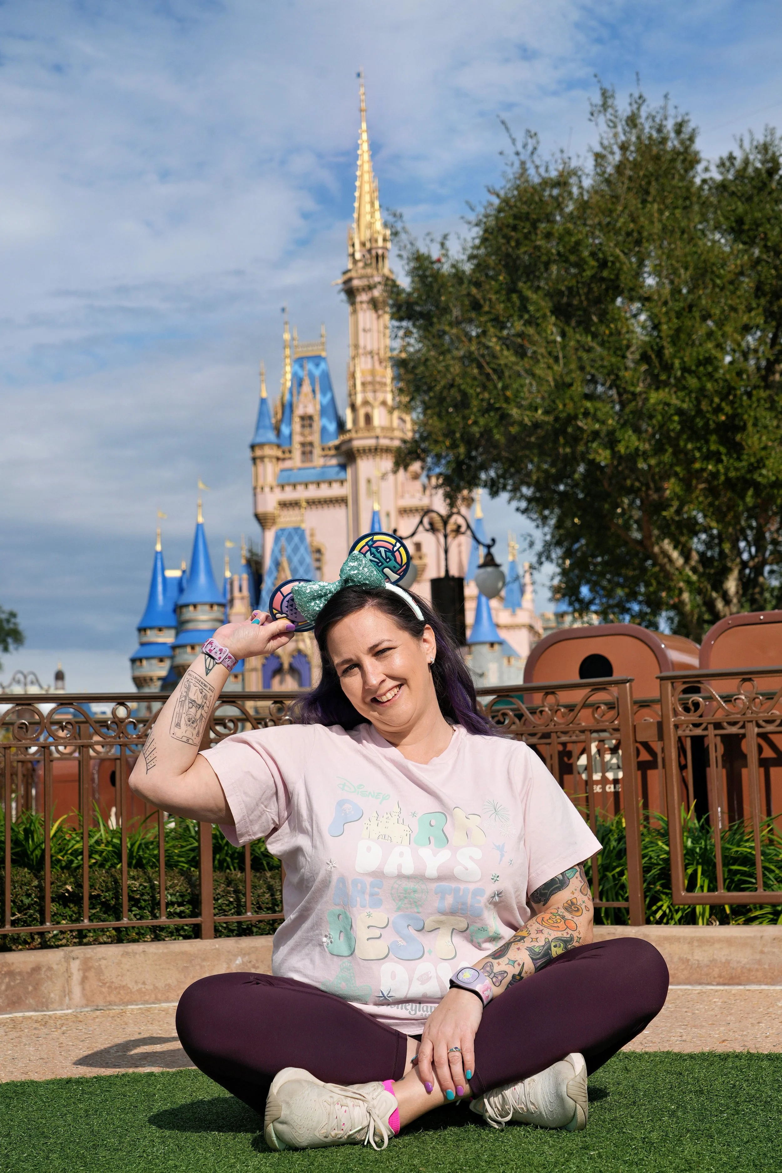 A woman with tattoos on her arms and purple hair, wearing a light pink Disney Pride Days shirt, sitting cross-legged on the grass in front of Cinderella Castle at Disney theme park. She is smiling, with one hand on her head and the other on her knee, wearing a green glittery bow headband with Minnie Mouse ears.