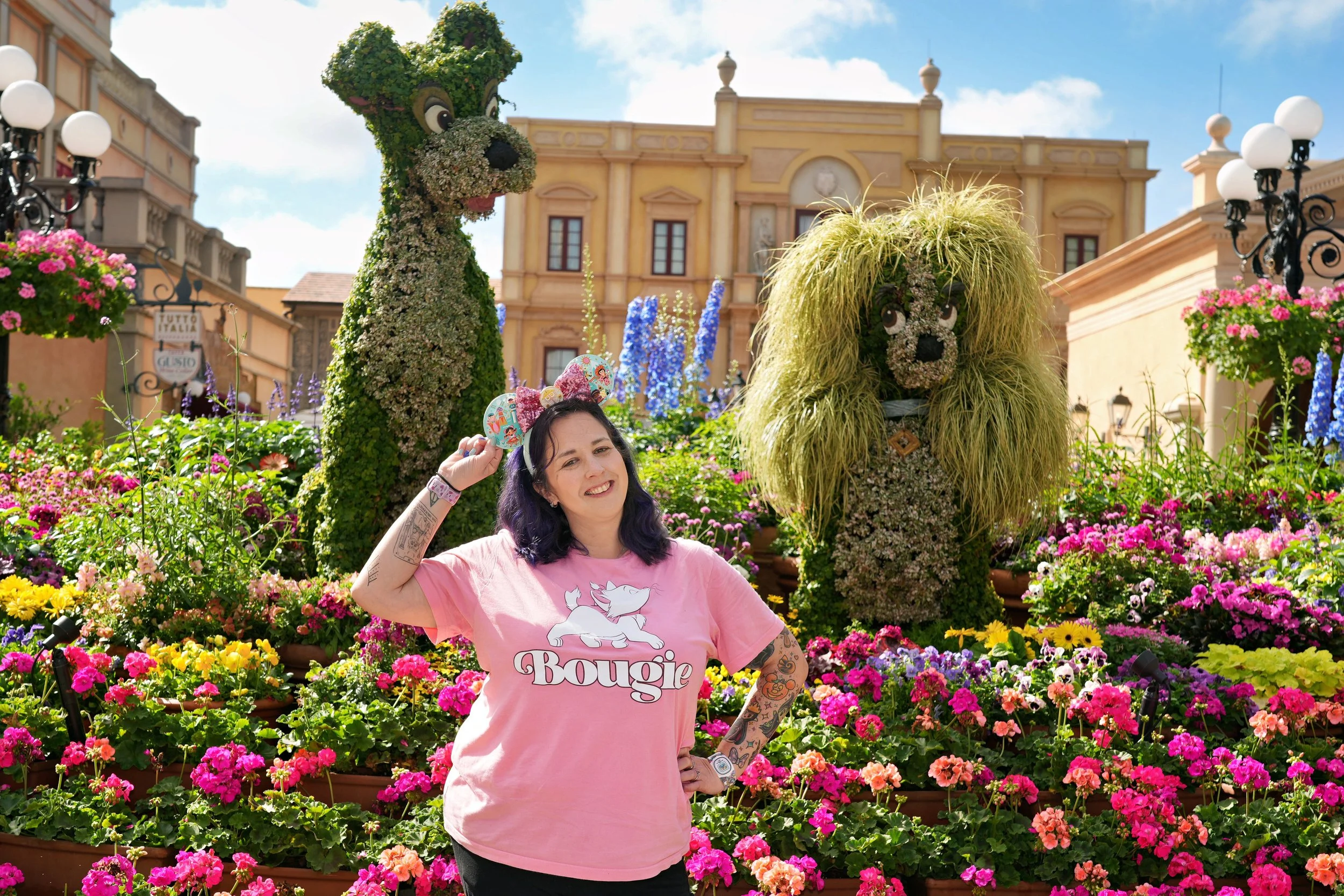 A woman standing in front of colorful flowers with two large topiary dogs behind her. She is smiling, wearing a pink t-shirt with a graphic of two dogs and the word 'Bowie', and adjusting Minnie Mouse ears on her head.