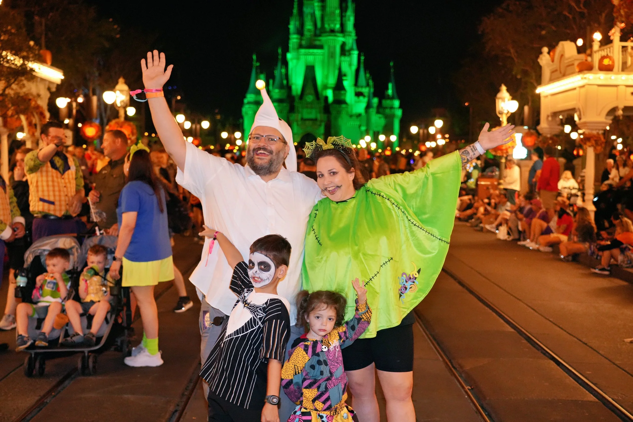 People in Halloween costumes enjoy a nighttime parade at a theme park, with a castle illuminated in green in the background.