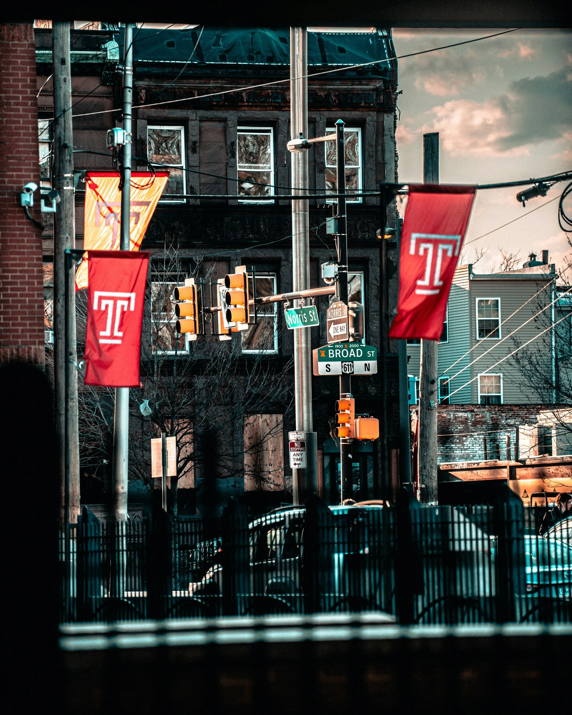 Street view with flags, traffic lights, and street signs at the intersection of Norris Street and Broad Street, Philadelphia, near Temple University, with buildings, cars, and power lines in the background.
