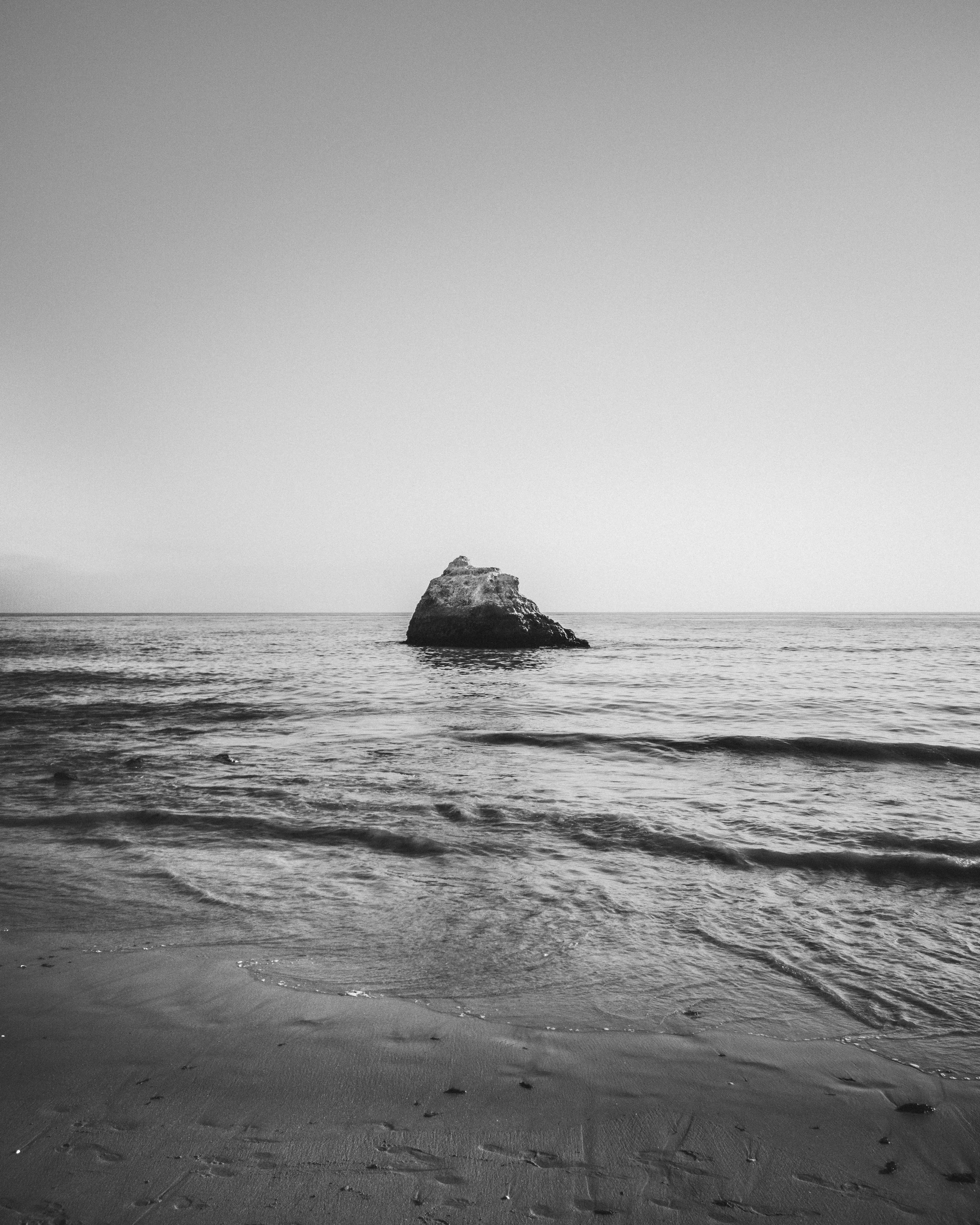 Black and white photo of a large rock in the ocean near the shoreline, with gentle waves and a vast sky.