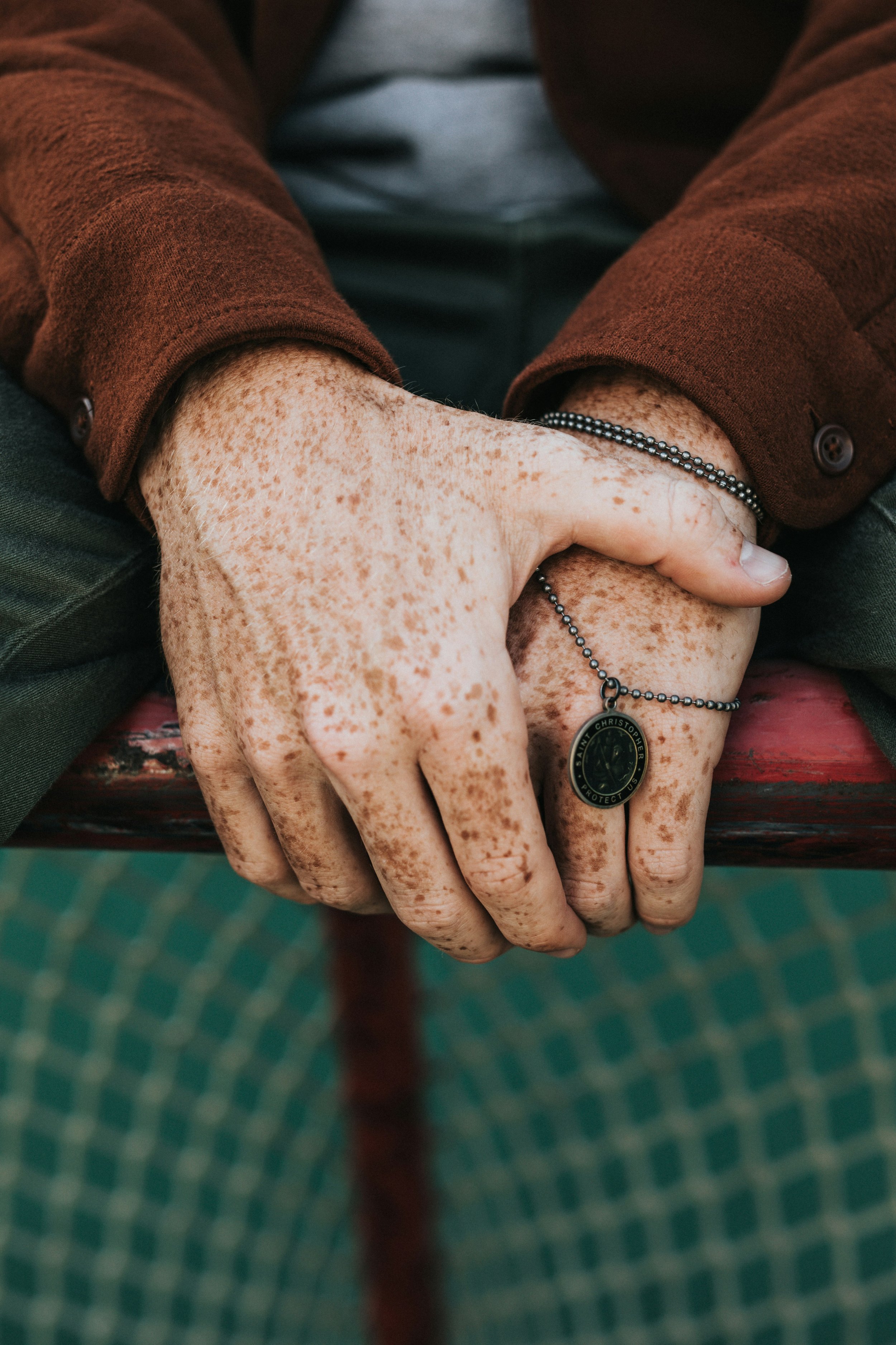 Close-up of a person's freckled hands resting on a red bench, with a beaded necklace hanging from one hand, wearing a brown long-sleeve shirt and green camouflage patterned pants.