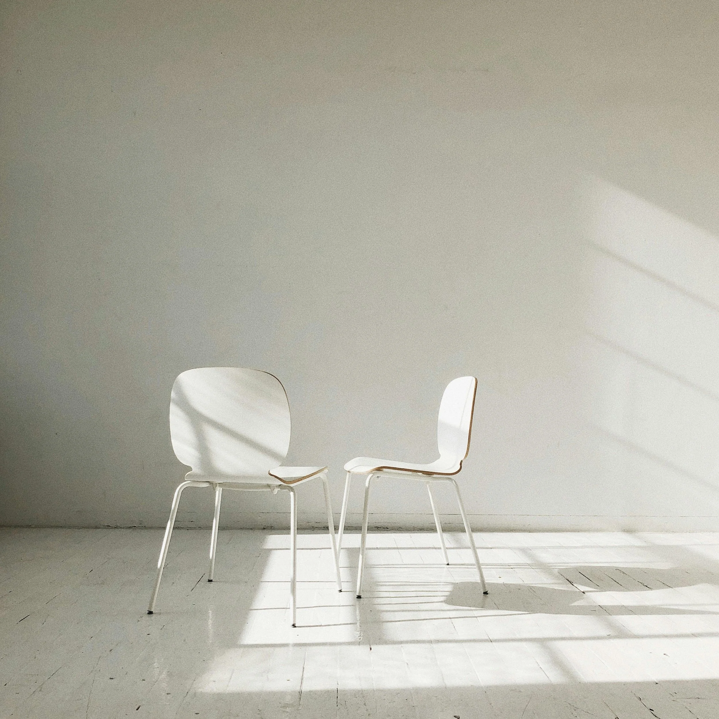 Two white chairs with curved backrests in a minimalistic room, casting shadows on a white wall and floor.