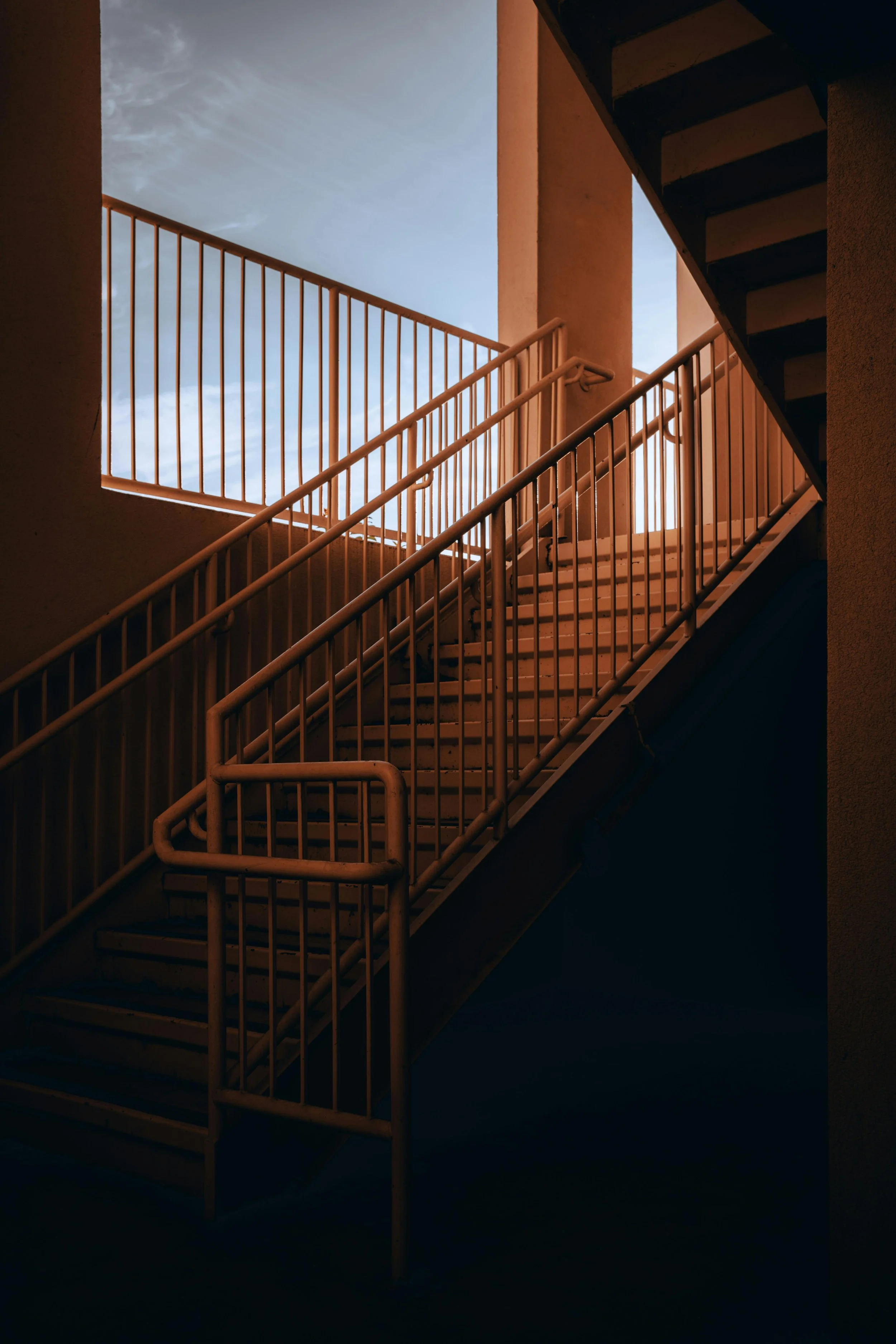 Exterior stairway with metal handrails leading upward, with the sky visible through an opening at the top of the stairs.