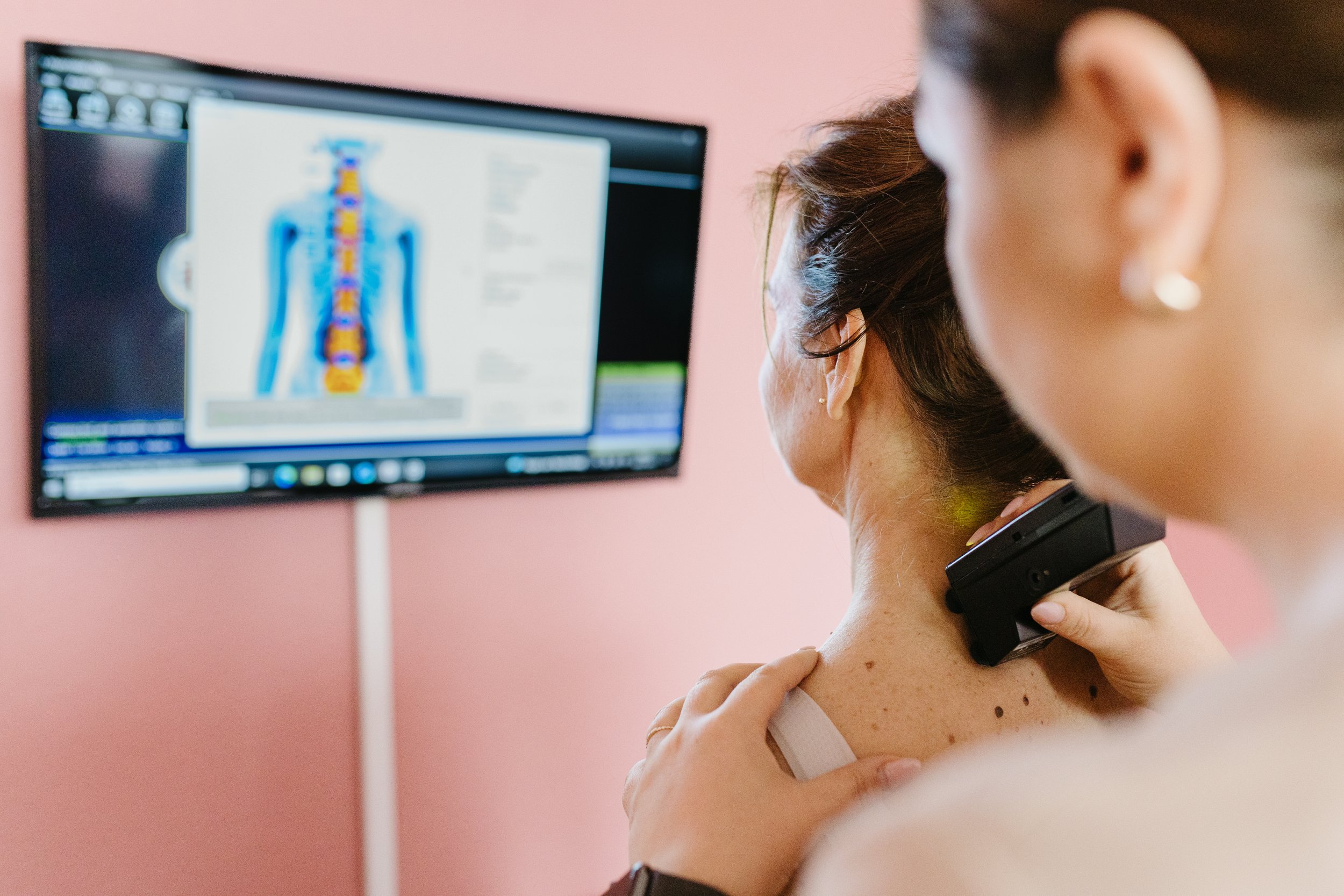 A woman is undergoing a digital scan or diagnostic procedure on her neck, with a healthcare professional holding a scanner or device on her neck and a monitor displaying a visual of the human body's spinal and nervous system in the background.