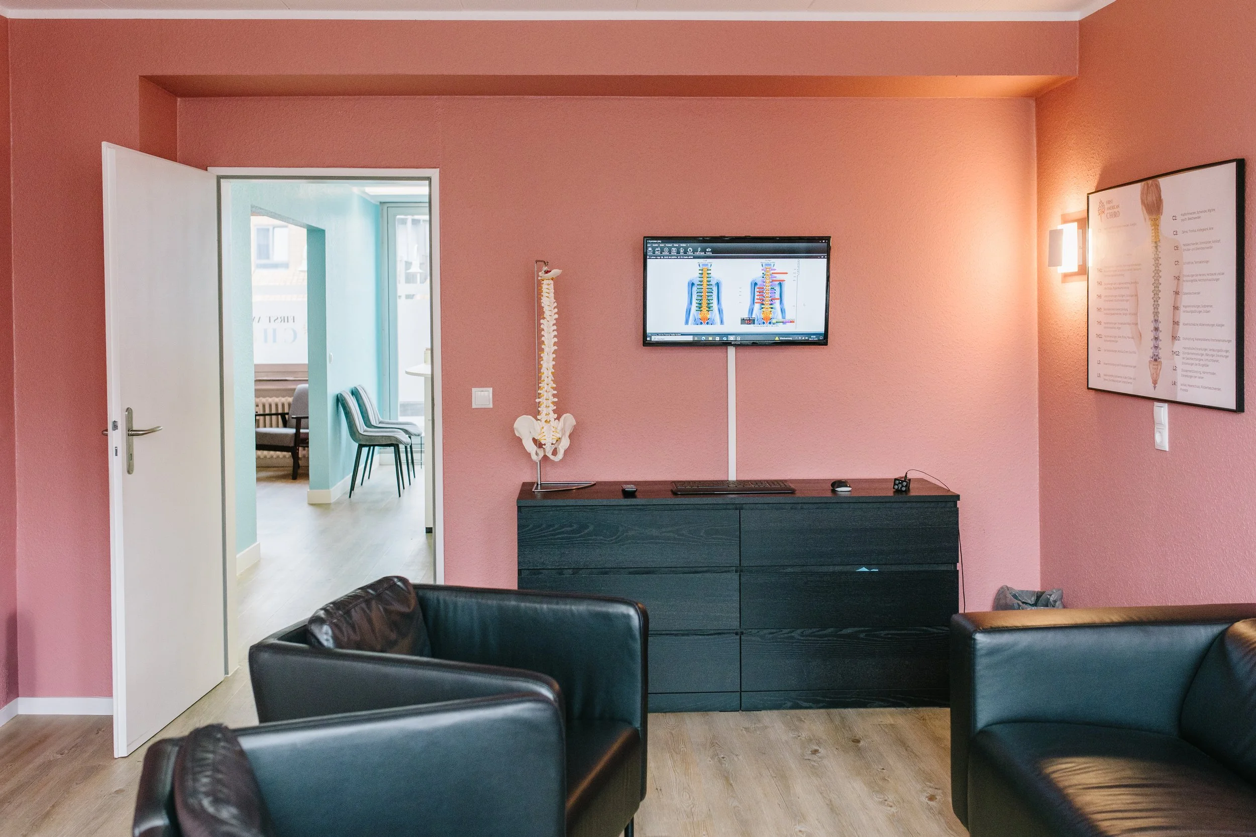 A waiting room with pink walls, black leather chairs, a black cabinet, a wall-mounted screen displaying a spine diagram, a skeleton model, and a large poster of a human spine on the wall.