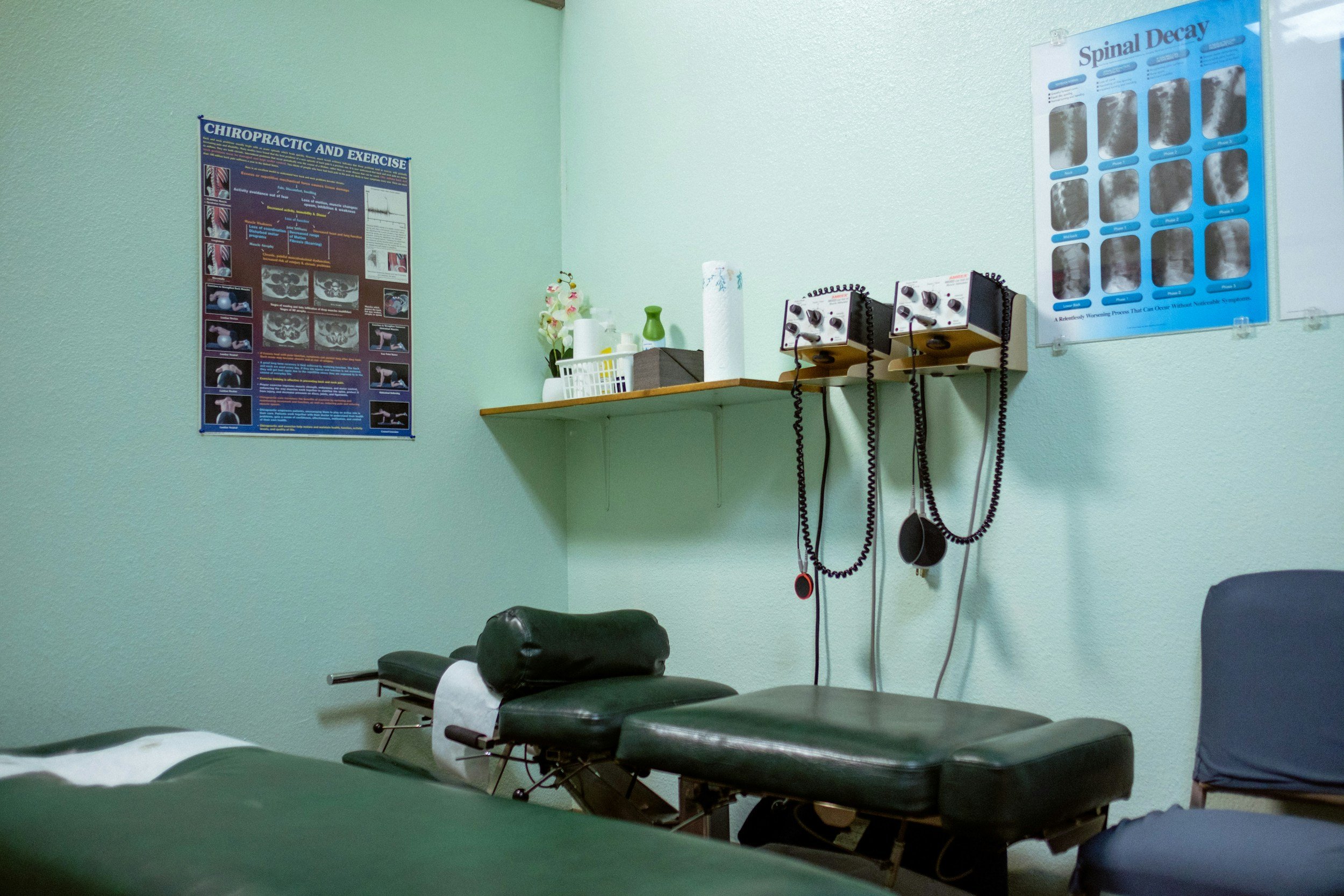 A medical examination room with chiropractic equipment, including an examination table, posters on the wall about spinal health, and a green chair.