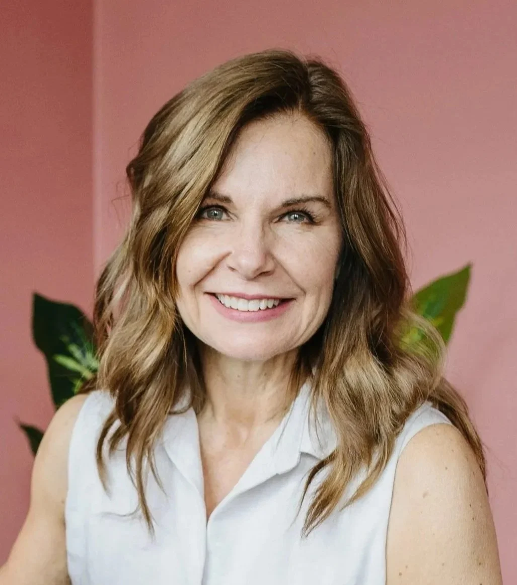 Smiling woman with wavy brown hair, wearing a sleeveless white shirt, against a pink background with green leaves.