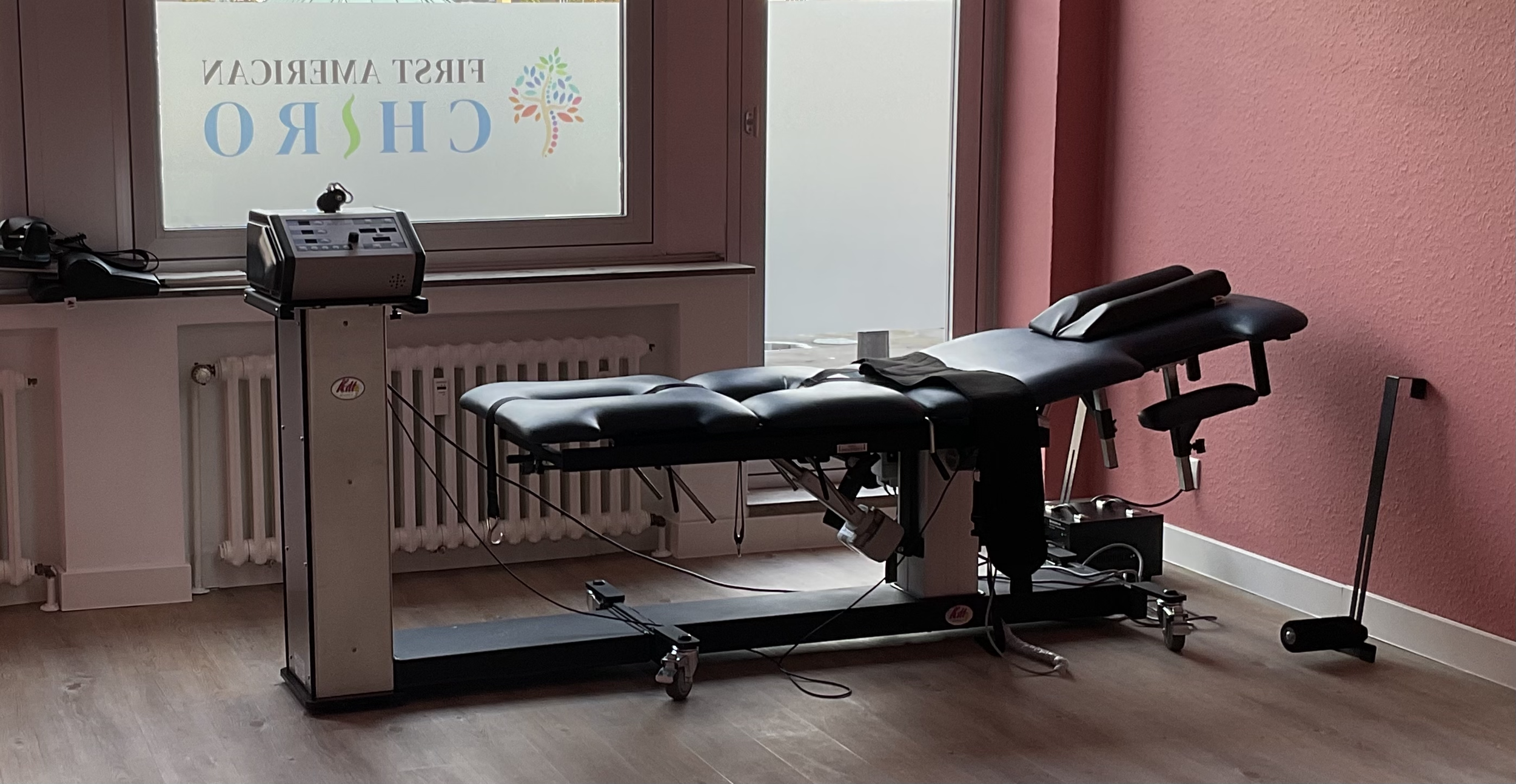 A chiropractic examination room with a chiropractic table, a small electronic device on a stand, and a window with the word 'CHIRO' visible through frosted glass, pink walls, and wood flooring.