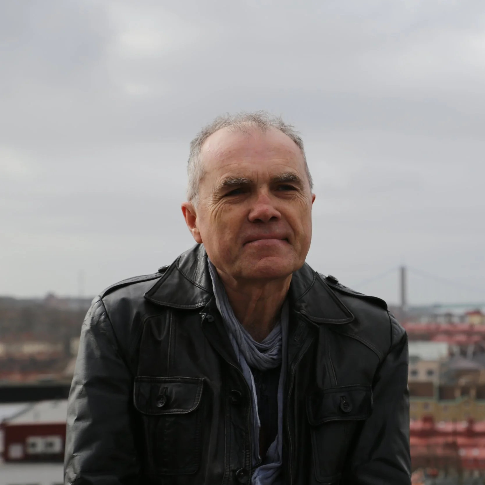 A middle-aged man with short gray hair smiling outdoors against a cloudy sky, wearing a black leather jacket over a striped shirt.