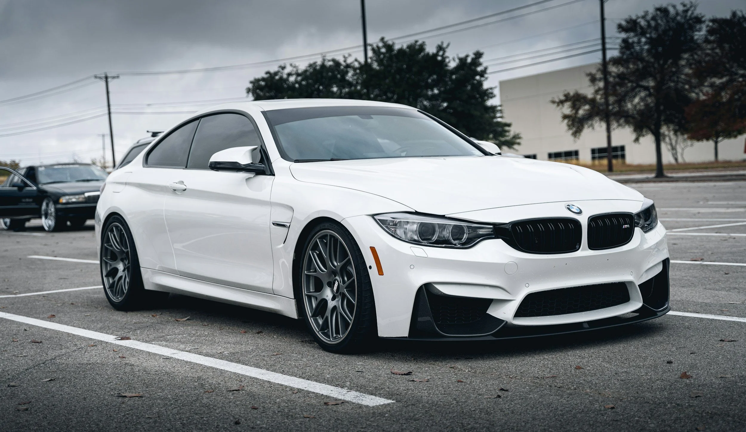 White BMW sports car parked in an empty lot on a cloudy day, with nearby trees and a building in the background.